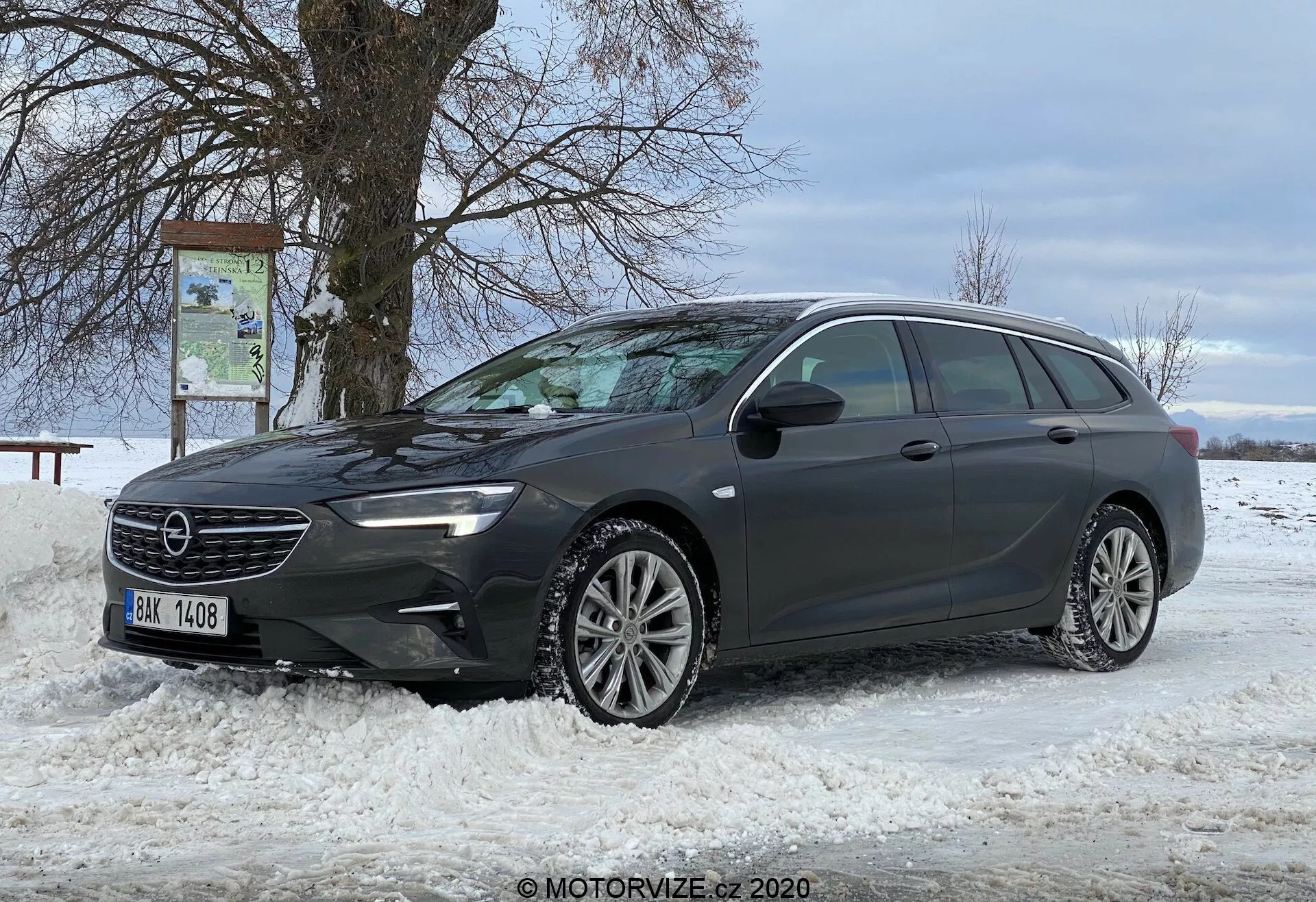 Cette image montre un break moderne, l'Opel Insignia Sports Tourer 2020, garé sur un sol enneigé. La voiture est positionnée en diagonale, offrant une vue claire de son profil avant et de tout le côté conducteur. L'angle de la prise de vue est une vue trois quarts avant, mettant en valeur la conception de la grille, les phares à LED élégants, le pare-chocs avant et les roues en alliage à multiples rayons. La longueur du break, la configuration des fenêtres et le système de barres de toit sont visibles, suggérant une utilité pour le transport de charges. Un rétroviseur latéral, probablement équipé de technologies avancées, et un fond de scène avec un arbre dénudé et un ciel nuageux ajoutent à la mise en scène.