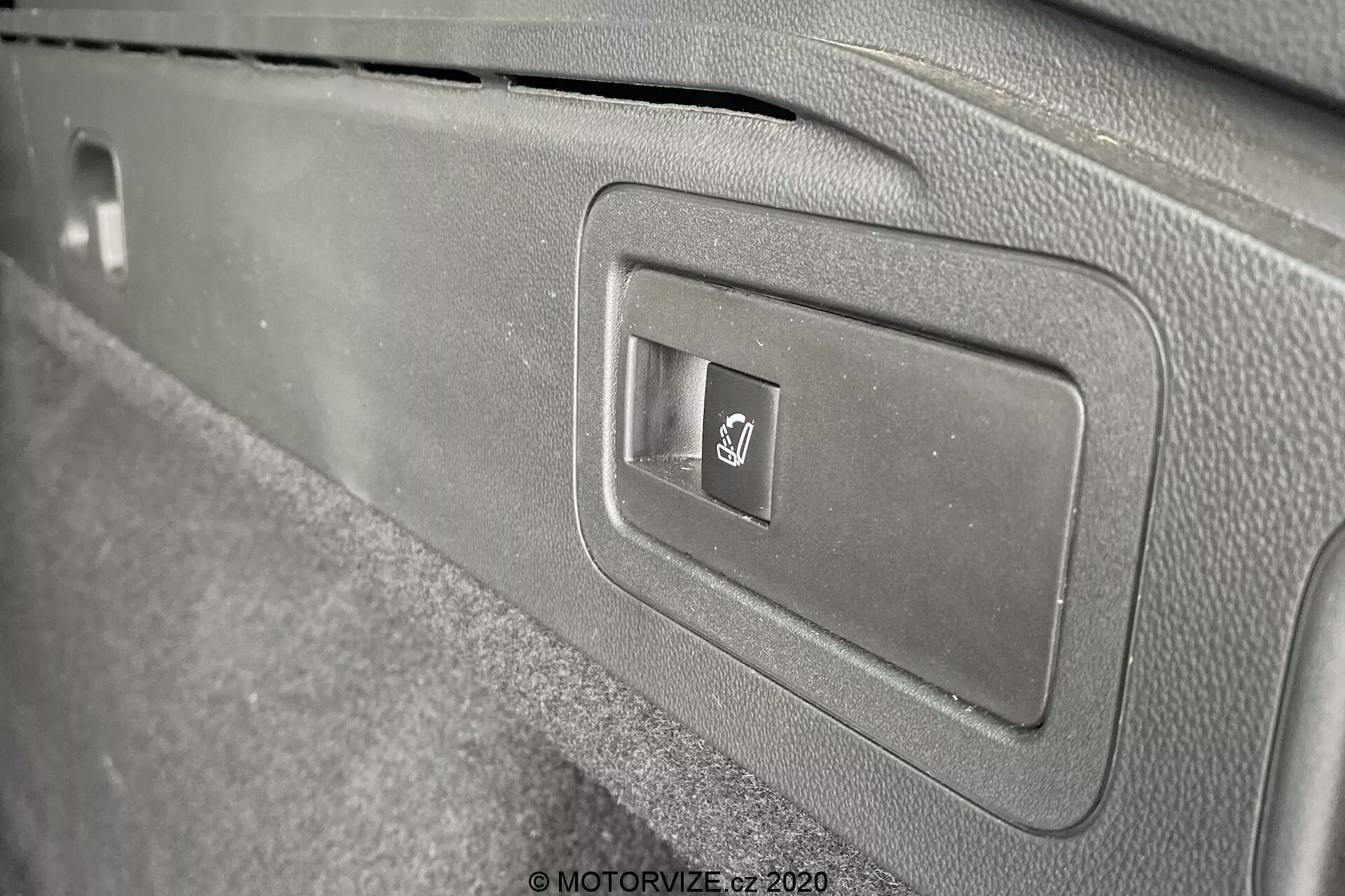 Gros plan du panneau intérieur de la Opel Insignia Sports Tourer 2020, mettant en avant un volet en plastique rectangulaire avec un bouton de déverrouillage du coffre électronique. Le bouton est marqué du symbole universel du coffre. L'image, prise sous un angle oblique, montre le design épuré du panneau avec ses différentes nuances de gris et une texture de type moquette autour du bouton.