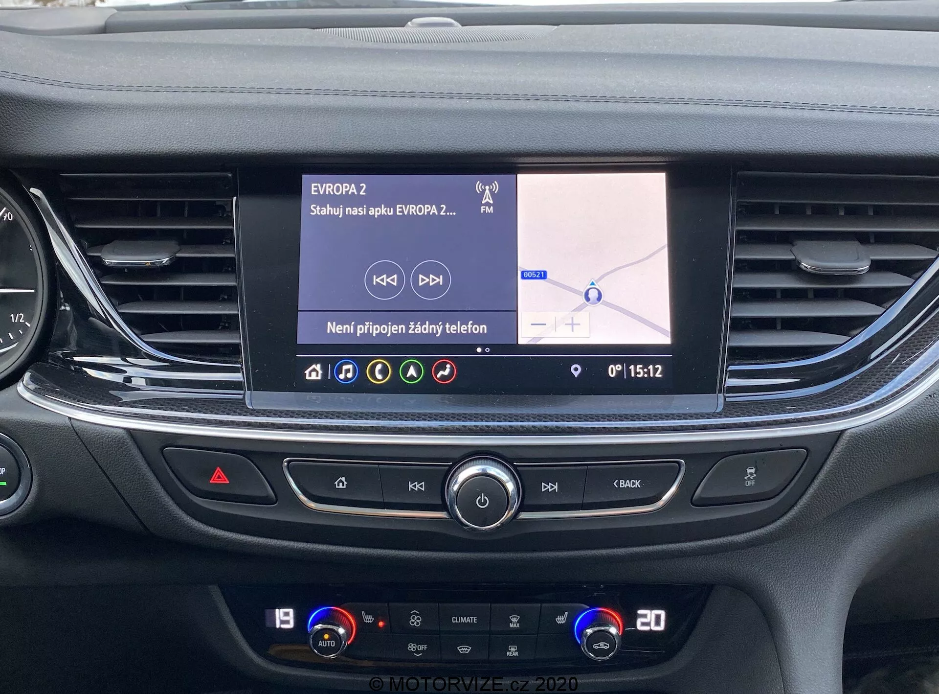 Vue de la console centrale de l'Opel Insignia 2020 depuis le siège conducteur ou passager avant, montrant le système d'infodivertissement et le panneau de commande de la climatisation. L'écran tactile LCD couleur, d'environ 7 à 10 pouces, affiche une carte de navigation avec du texte en langue slave et l'étiquette 'EVROPA2', suggérant une position en Europe de l'Est ou Centrale. L'interface indique qu'aucun téléphone n'est connecté et l'heure est 15:12. En dessous, on trouve des bouches d'aération horizontales et des boutons pour le contrôle multimédia, les feux de détresse et d'autres réglages, suivis du système de contrôle du climat à deux zones indiquant 19 et 20 degrés Celsius. Le design élégant et moderne met en avant la technologie de pointe de la voiture pour une expérience utilisateur pratique.