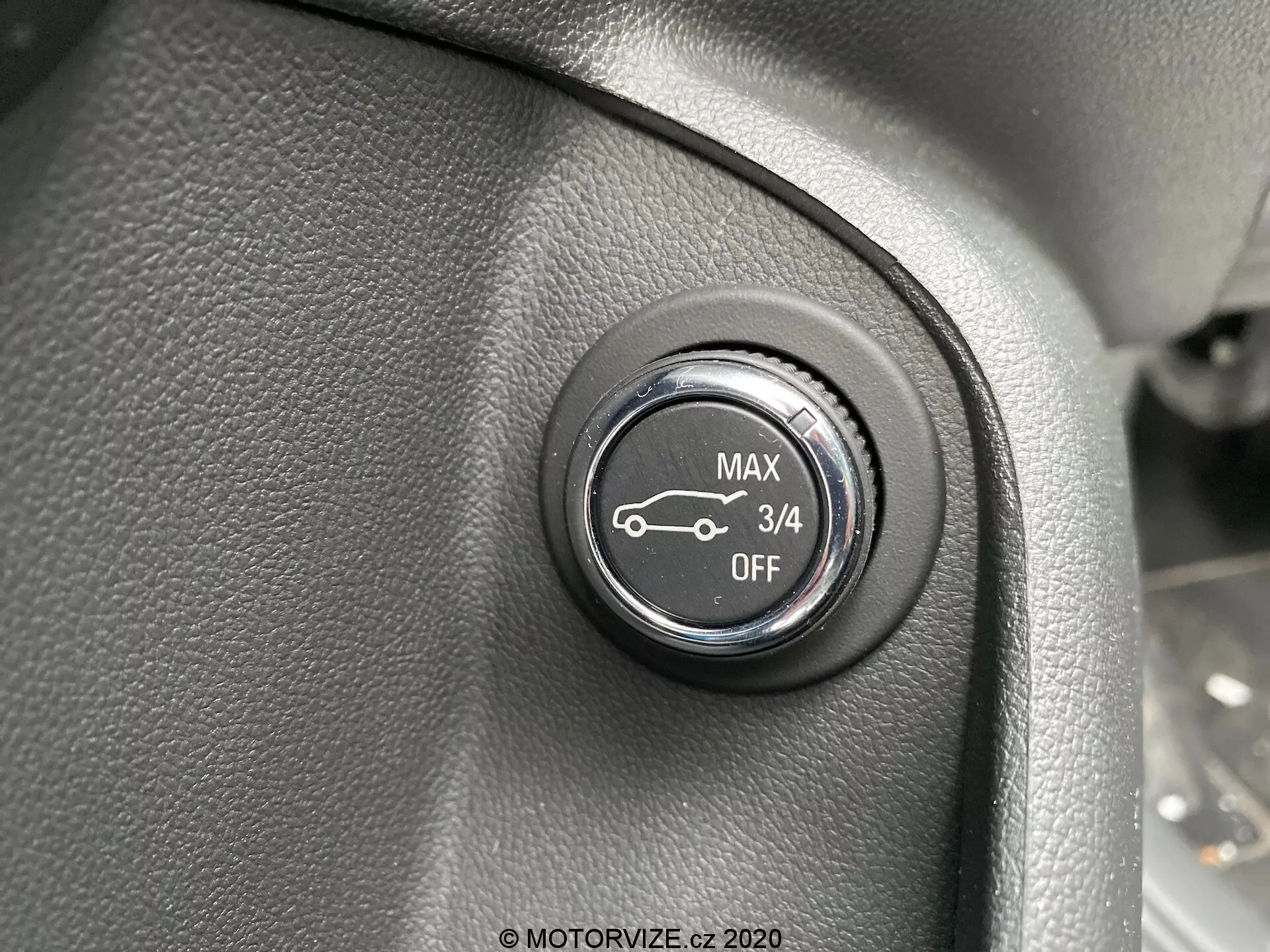 Vue rapprochée d'un bouton de contrôle à l'intérieur de l'Opel Insignia 2020. Positionné sur une surface texturée, probablement une partie du tableau de bord ou du panneau latéral près du volant, le bouton noir avec des marquages blancs présente quatre positions: 'MAX,' un symbole de voiture avec une flèche vers le haut, '3/4' et 'OFF.' Cela suggère qu'il contrôle une fonction telle que le réglage des phares ou des suspensions. La photo se concentre sur la face du bouton, légèrement au-dessus, avec une faible profondeur de champ et un éclairage uniforme.