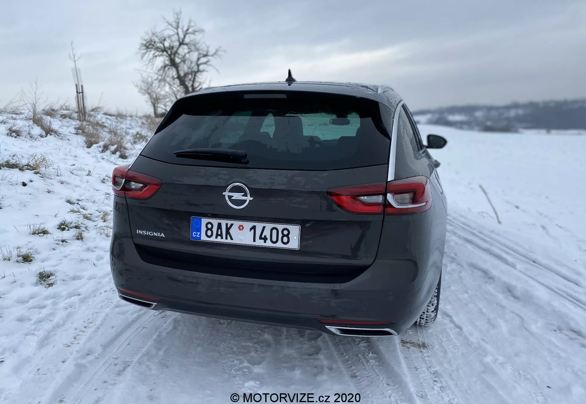 Vue arrière de l'élégante et moderne Opel Insignia 2020 Sports Tourer dans un paysage enneigé, avec des feux arrière à LED étroits, un double échappement, une lunette arrière avec dégivreur, un troisième feu stop intégré dans un spoiler et une antenne aileron de requin sur le toit. L'image est prise d'un angle bas centré sur le côté gauche, mettant en évidence l'insigne et l'écusson du fabricant sur le coffre.