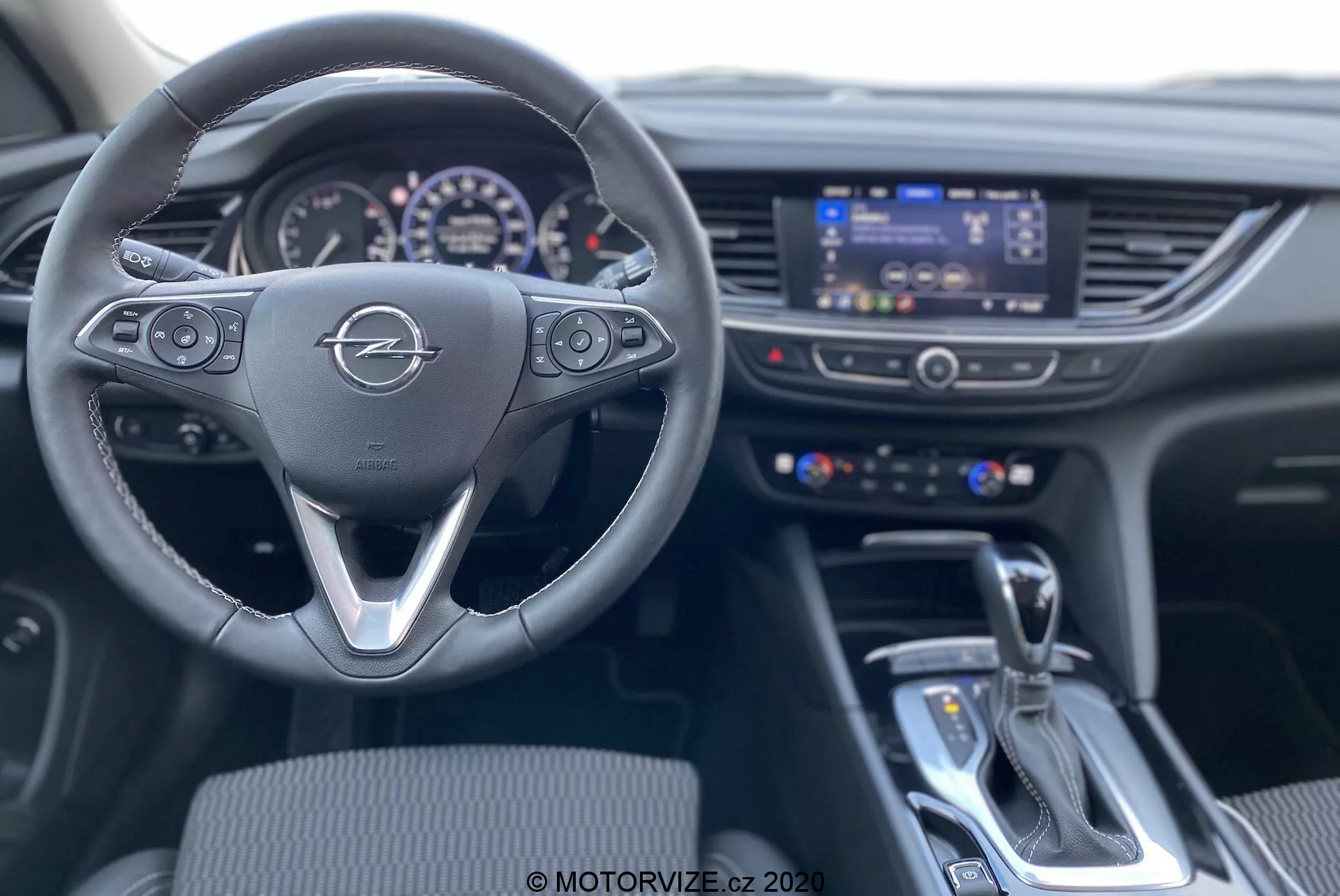 Vue intérieure depuis le siège du conducteur d'une Opel Insignia Sports Tourer 2020, centrée sur le volant à trois branches avec des boutons de commande multifonctions, le tableau de bord éclairé et l'écran central d'infodivertissement entouré d'interrupteurs de contrôle de la climatisation. L'image montre également le levier de vitesses pour la transmission automatique et l'élément de contrôle à proximité sur la console centrale. L'intérieur est moderne, mettant en avant la fonctionnalité et l'intégration technologique.
