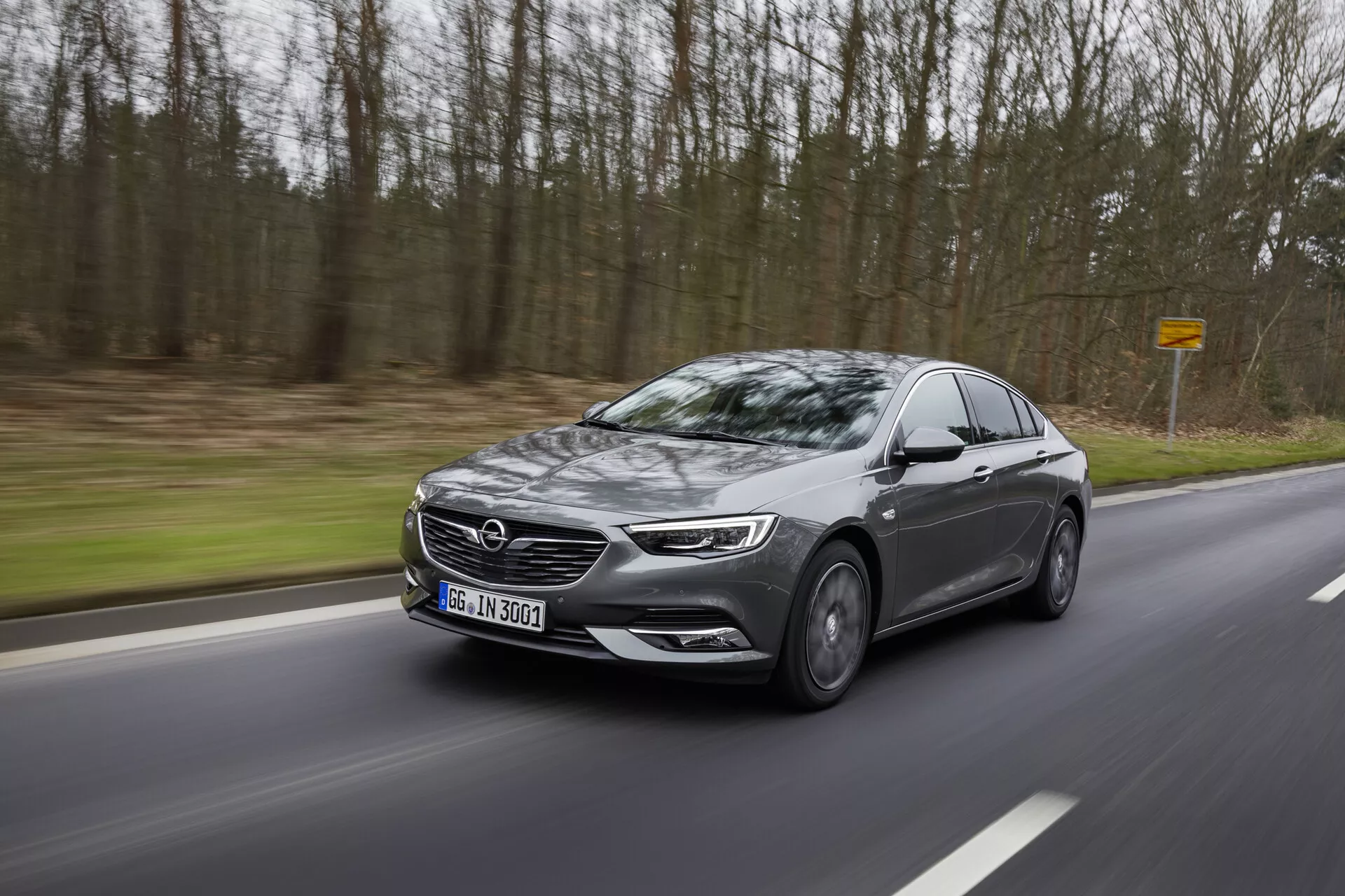 Sedan moderne Opel Insignia 2013 en mouvement sur une route sinueuse à travers les bois, affichant un profil latéral avec phares LED, grande calandre, roues en alliage, rétroviseurs latéraux avec indicateurs et vitres teintées