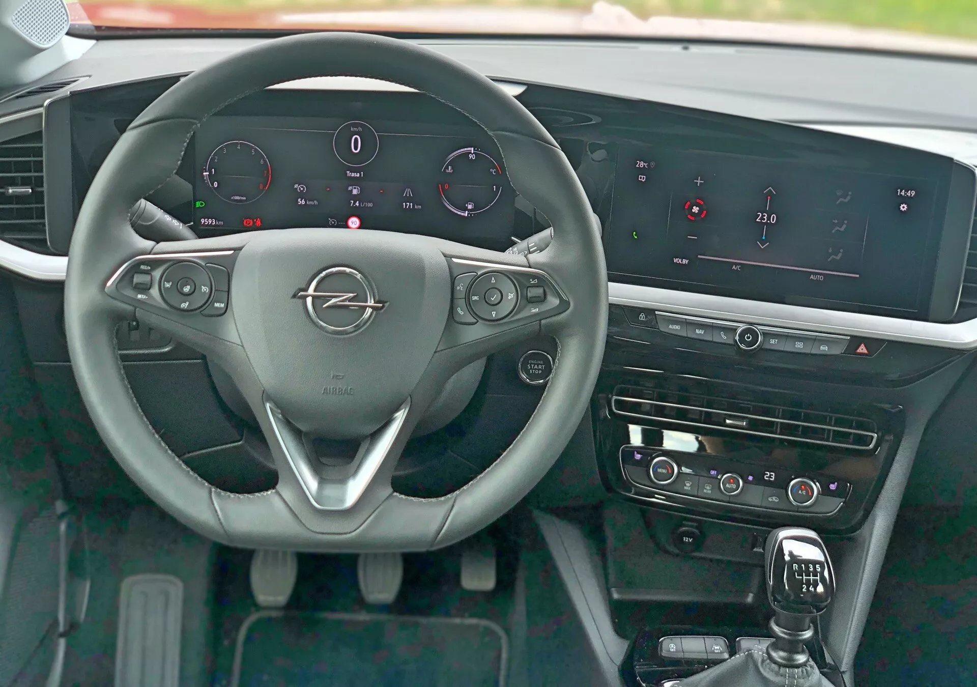 Vue intérieure de l'Opel Mokka 2021 depuis la perspective du siège conducteur montrant le volant à trois branches avec emblème métallique de la marque, commandes au volant pour audio, téléphone et régulateur de vitesse, palettes de changement, tableau de bord numérique affichant la vitesse, les RPM, le niveau de carburant et la température du moteur, système d'infodivertissement proéminent avec écran horizontal large, commandes tactiles pour HVAC, levier de changement de vitesse avec housse en cuir, éléments de garniture polis et bouton de démarrage sans clé.
