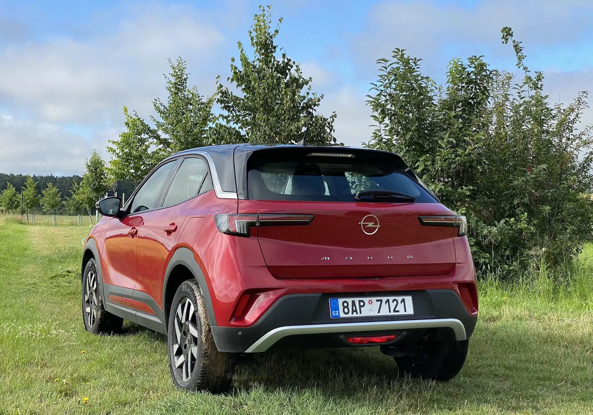 Photographie couleur montrant une vue trois quarts arrière d'un SUV crossover Opel Mokka 2021 moderne en rouge et noir, garé en extérieur dans une zone herbeuse avec des arbres ou des buissons en arrière-plan. Les éléments de design du véhicule comprennent un spoiler arrière aérodynamique, des feux arrière à LED angulaires, une lampe stop montée en hauteur, une carrosserie bicolore, de grandes roues en alliage, des poignées de porte affleurantes et des garnitures en plastique noir. La photo est prise d'un angle bas, mettant en évidence la hauteur et la garde au sol du SUV, avec un ciel partiellement nuageux indiquant des conditions météorologiques variables.