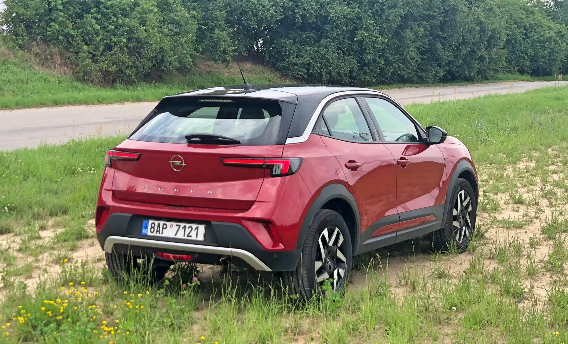 SUV crossover moderne Opel Mokka 2021 vu sous un angle trois quarts arrière, soulignant le spoiler arrière, le pare-brise arrière incliné, le badge de la marque clairement visible et les feux arrière élégants. Positionné sur une surface herbeuse, montrant son potentiel tout-terrain léger. Le profil latéral révèle des lignes sculptées de carrosserie, des protections inférieures noires et des roues au motif multi-branches stylé. Essuie-glace arrière intégré, capteurs de stationnement et pare-boue contrasté suggèrent aérodynamisme, praticité et robustesse.