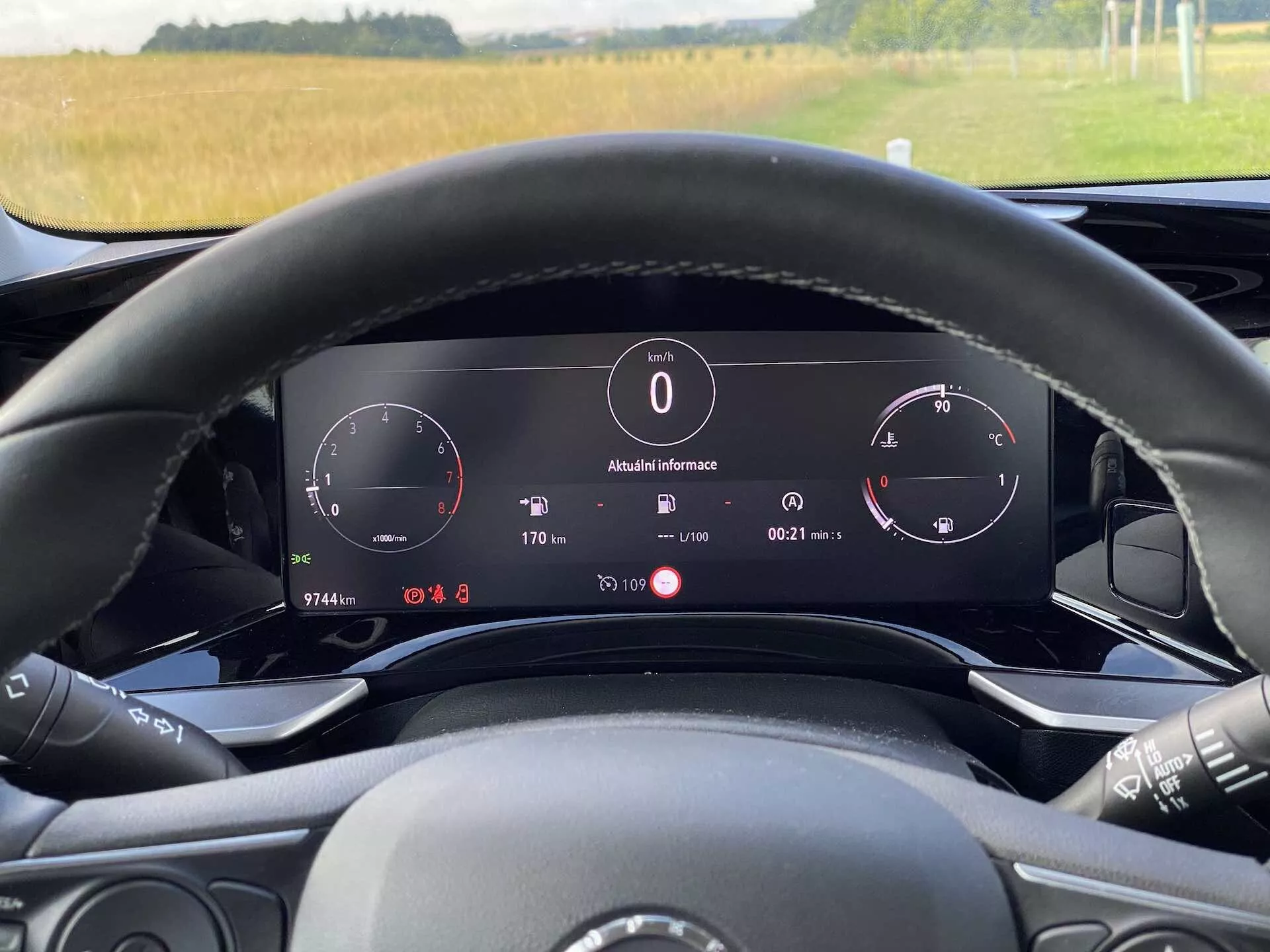 Vue intérieure de l'Opel Mokka 1.2 DI Turbo 2021 depuis la perspective du conducteur au volant. L'image met en avant le tableau de bord numérique derrière le volant incluant un compteur de vitesse, un compte-tours et un panneau d'informations affichant la température extérieure (8°C), l'heure, la position du rapport ('P' pour parking) et les détails de l'ordinateur de bord. Le volant avec les contrôles multimédias est partiellement visible. L'éclairage ambiant suggère une scène de jour avec une vue rurale à travers le pare-brise. Le véhicule présente un tableau de bord à affichage électronique et des fonctionnalités technologiques avancées.