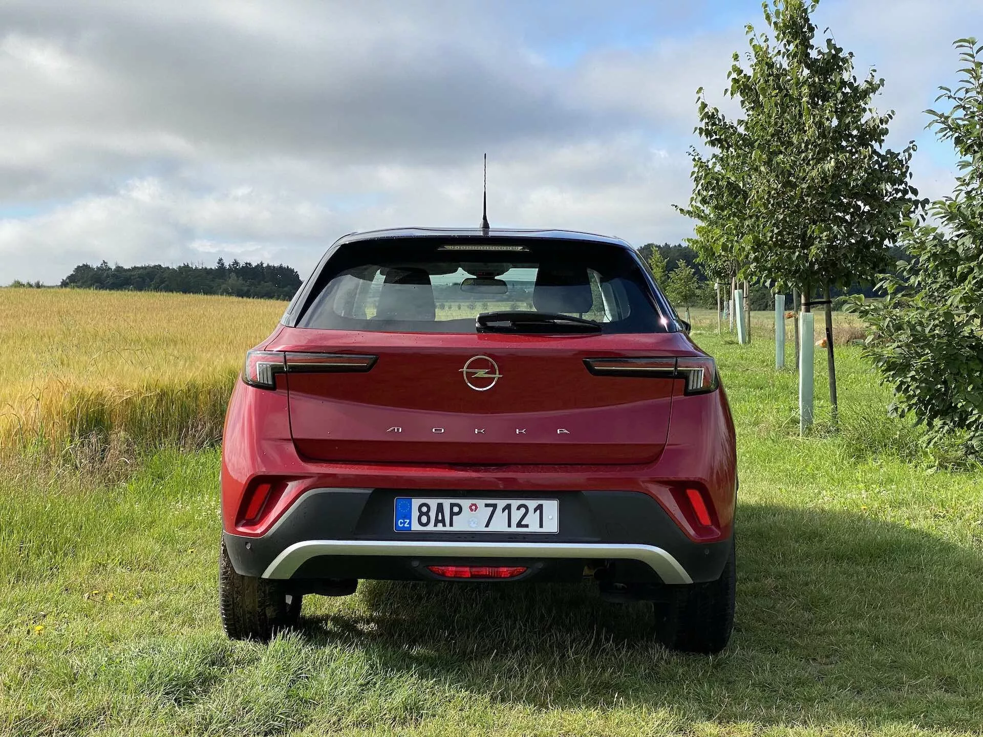 Vue arrière d'une Opel Mokka 1.2 DI Turbo 2021 rouge, présentant un design moderne avec des lignes acérées et l'emblème du fabricant sur le hayon, des clusters de feux arrière distinctifs à LED, un pare-chocs arrière avec inserts contrastants, des capteurs de stationnement, des réflecteurs intégrés, un essuie-glace arrière, une antenne en forme d'aileron de requin, un becquet arrière et une plaque d'immatriculation dans un cadre rural avec de l'herbe, un champ de culture et des arbres.