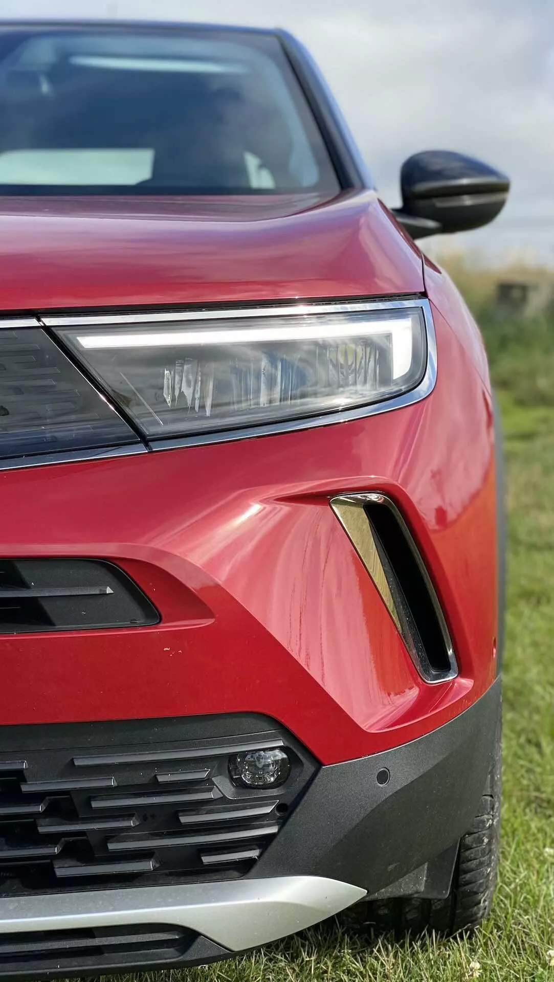 Vue rapprochée et angulaire du côté avant gauche d'une Opel Mokka 2021 rouge. La photo capture le phare avec éclairage LED ou HID, une partie de la grille avec des lamelles horizontales, le logement du feu antibrouillard avec un contour noir et argent, une peinture de carrosserie rouge vibrante avec des contours anguleux audacieux, le rétroviseur aérodynamique et le capot et l'aile partiellement visibles. La voiture est garée sur l'herbe avec un arrière-plan flou.