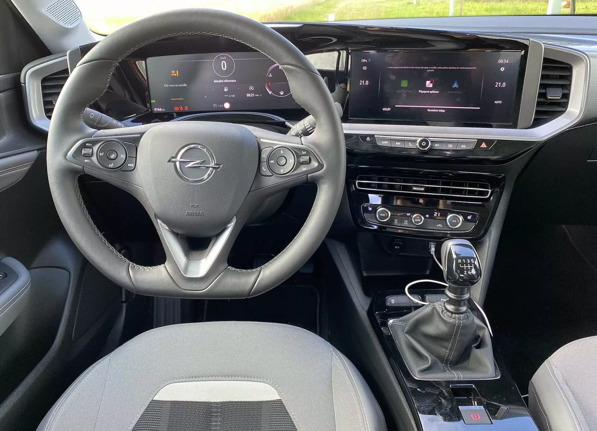 Intérieur de l'Opel Mokka 2021 depuis la perspective du passager avant, avec un volant à trois branches avec commandes intégrées, un tableau de bord entièrement numérique, un écran tactile central d'infodivertissement avec contrôle climatique et navigation et un levier de vitesse manuel. La disposition comprend des systèmes de contrôle doubles et des boutons supplémentaires pour les modes de conduite, avec des sièges en tissu.