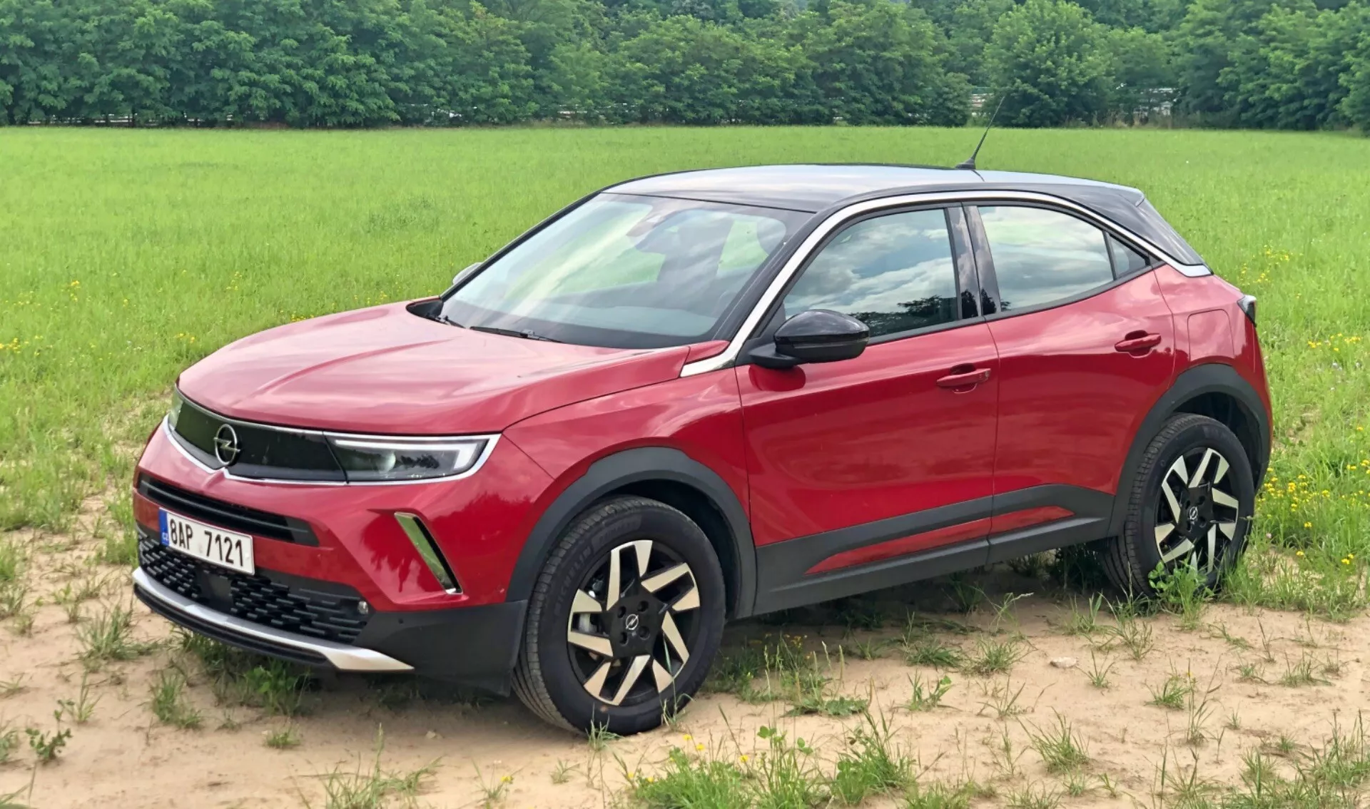 Image d'un SUV Opel Mokka 2021 rouge garé sur une surface herbeuse avec un champ en arrière-plan. Cette vue trois quarts avant met en évidence la calandre, potentiellement indiquant une technologie de groupe motopropulseur avancée, des feux de jour à LED élégants, les feux antibrouillard et des jantes en alliage bicolores uniques. Les rétroviseurs et poignées de porte de couleur carrosserie, la ligne d'épaule dynamique, la garniture inférieure noire protectrice et les cadres de fenêtre chromés sont également visibles, montrant les caractéristiques de design modernes et luxueuses du véhicule.