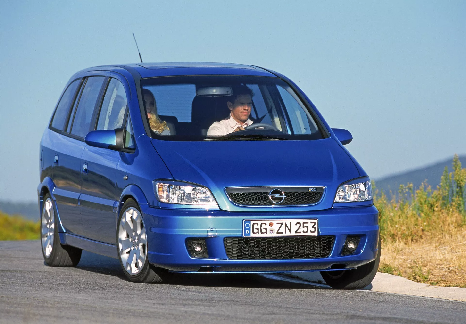 Image d’un Opel Zafira (A-facelift) compact MPV bleu de 2002 en vue trois quarts avant basse, mettant en avant la face avant de la voiture avec une visibilité claire de la calandre, des phares halogènes avec clignotants intégrés, pare-chocs avant avec prise d’air et bouches d’aération latérales auxiliaires, capot aérodynamique avec des contours subtils, grandes roues en alliage multiplis et un court porte-à-faux avant suggérant une disposition à traction avant. Un conducteur avec ceinture de sécurité est visible à travers le pare-brise avant, soulignant les caractéristiques de sécurité routière.
