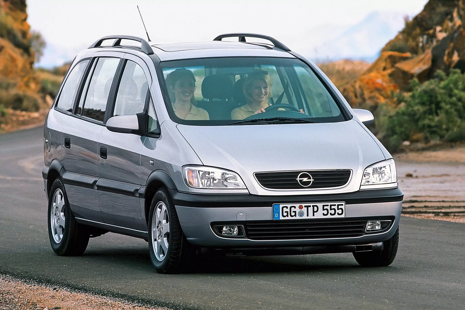 Photographie d'un Opel Zafira de 1999, un MPV compact, prise d'une perspective frontale trois quarts basse. L'image met en avant la calandre de la voiture avec un badge central, les grands phares probablement à sources lumineuses halogènes, le capot lisse, le pare-chocs avant avec antibrouillards, les jantes en alliage et le côté passager du véhicule avec une antenne à l'arrière du toit. Le véhicule est sur une route asphaltée avec un paysage naturel en arrière-plan. À travers le pare-brise avant, deux occupants sont visibles, reflétant la conception intérieure et l'espace du véhicule.