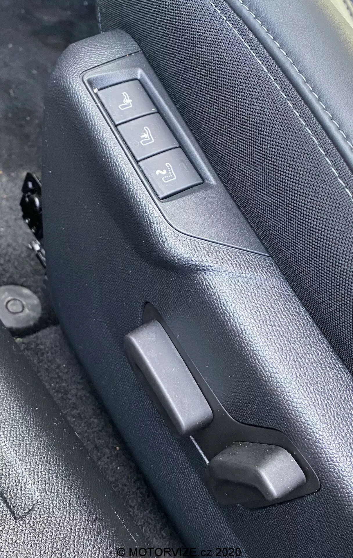 Gros plan des commandes de siège intérieur de la Peugeot 2008 1.2 PureTech 100 Active (2020), mettant en valeur le côté d'un siège en cuir noir ou gris foncé avec des coutures détaillées, vu sous un angle oblique légèrement surélevé. Le siège comporte trois boutons de réglage électronique et un levier manuel. Les boutons contrôlent les mouvements avant/arrière, l'inclinaison du dossier et la hauteur totale du siège, montés sur un panneau texturé assorti. Le filigrane indique 'MOTORVIZE.cz 2020'.