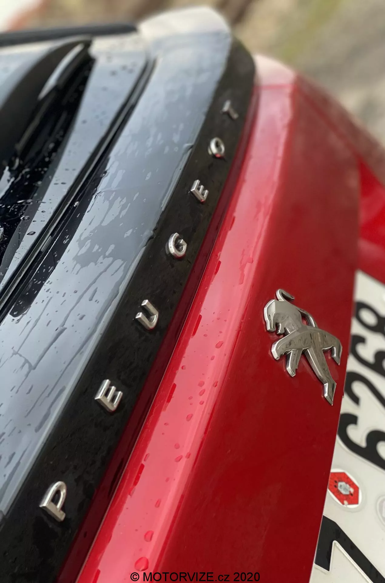 Gros plan sur l'emblème arrière et la désignation du modèle Peugeot 2008 2020 (1.2 PureTech 100 Active). La prise de vue est à un angle diagonal peu conventionnel, capturant le logo Peugeot en chrome sur un panneau de carrosserie rouge vif. Des lettres chromées espacées à gauche du logo forment 'PEUGEOT'. Une surface noire brillante avec des gouttelettes d'eau apparaît au-dessus du nom, indiquant une pluie récente ou un lavage de voiture. La section de plaque d'immatriculation floue est visible en bas à droite. La profondeur de champ réduite met en valeur l'emblème et le nom, tandis que d'autres éléments sont flous pour une touche artistique.