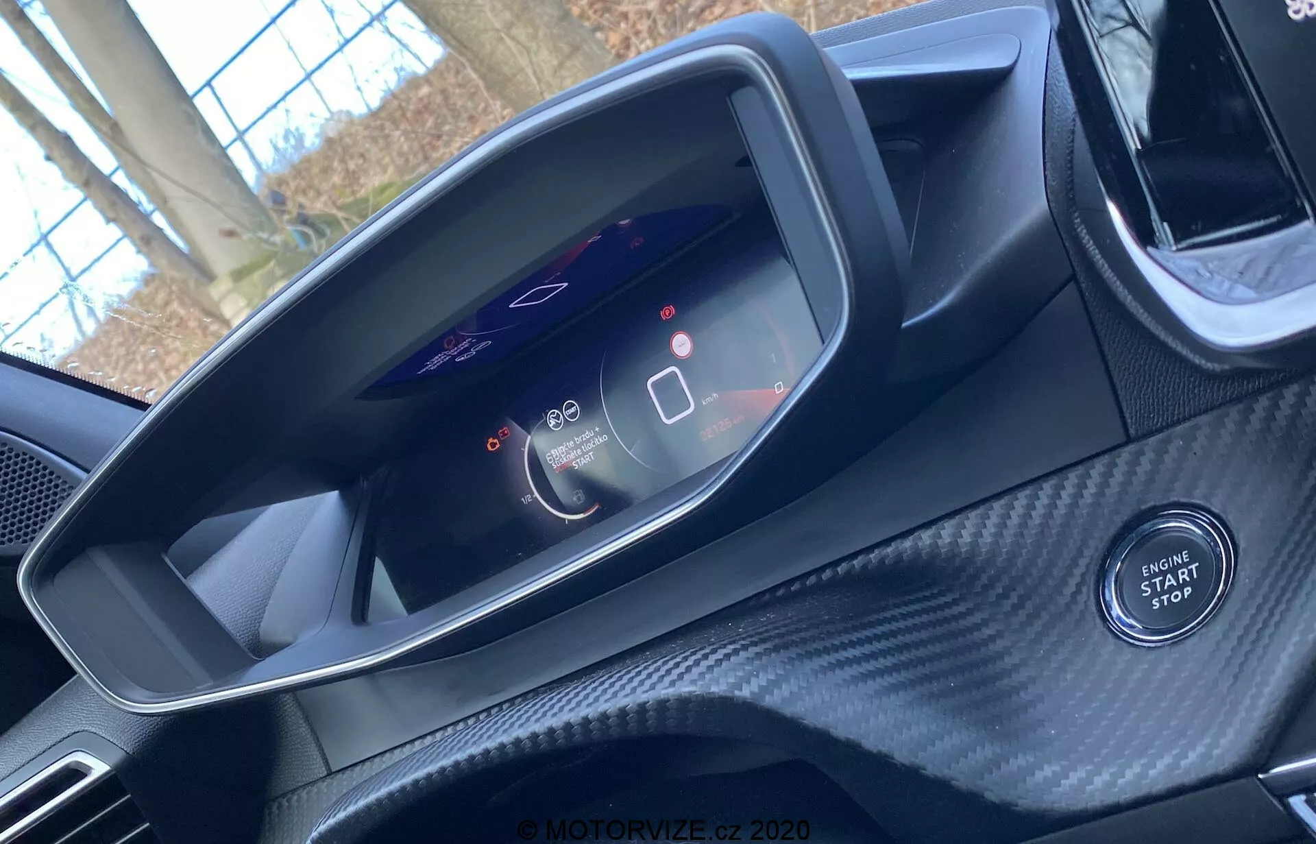 Vue intérieure d'une Peugeot 2008 2020 depuis la perspective du siège conducteur. L'image se concentre sur le tableau de bord numérique moderne affichant un compteur de vitesse à 0 et une jauge de consommation d'énergie. Elle montre également des icônes de systèmes avancés d'assistance à la conduite. Le bouton de démarrage/arrêt du moteur est visible sous le tableau de bord, indiquant l'allumage sans clé de la voiture. La zone environnante présente une texture semblable à la fibre de carbone, suggérant un intérieur sportif ou haut de gamme. Le haut du volant est à peine visible.