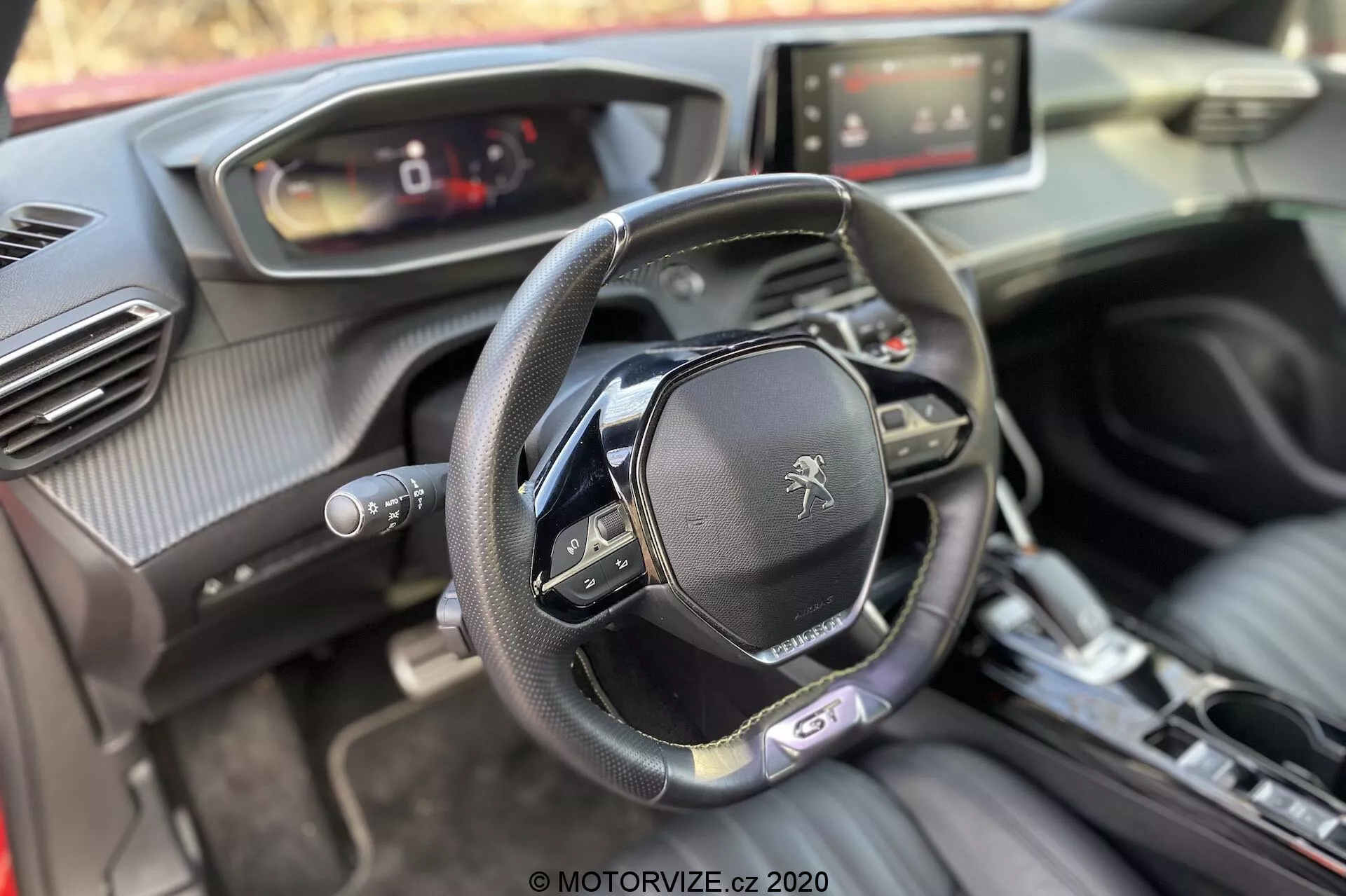 Intérieur de la Peugeot 2008 1.2 PureTech 100 Active de 2020 vu depuis la perspective du passager avant, centré sur le cockpit du conducteur. L'image montre un volant à fond plat avec plusieurs boutons de contrôle et le logo lion de Peugeot, un tableau de bord numérique, un système d'infodivertissement à écran tactile, une console centrale de transmission automatique, un bouton de démarrage/arrêt, des aérations avec des accents chromés et un mélange de matériaux doux au toucher, y compris du cuir et des textures similaires à la fibre de carbone.