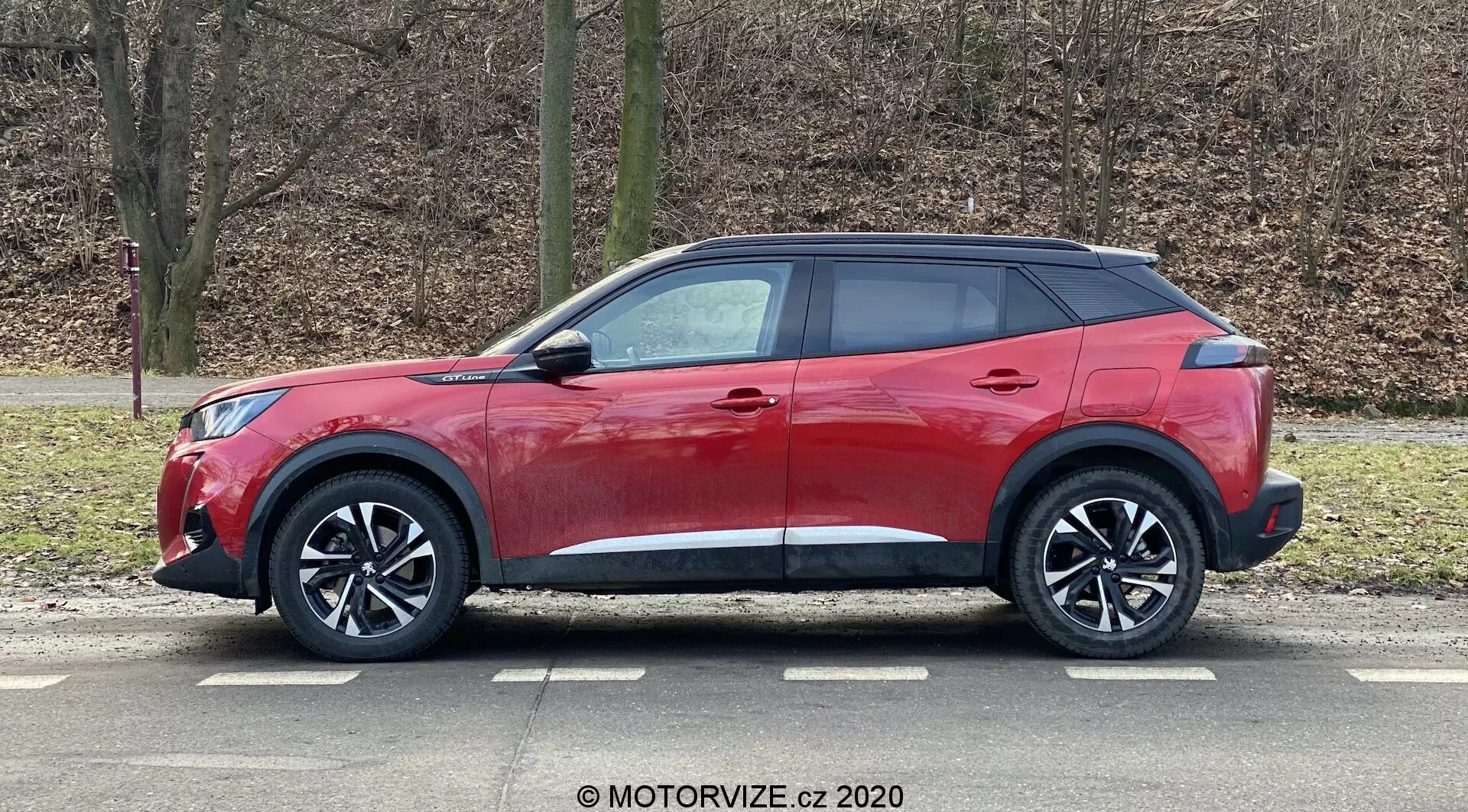 Véhicule utilitaire sport (SUV) Peugeot 2008 2020 1.2 PureTech 100 Active rouge avec un design moderne et élégant, ligne de toit inclinée, ceinture de caisse haute avec fenêtres se rétrécissant, poignées de porte aérodynamiques, rétroviseurs latéraux avec clignotants intégrés, grandes roues alliage multi-rayons, pneus à profil bas, moulures en plastique noir pour une apparence robuste, peinture rouge brillante et vitres arrière teintées, capturé en vue de profil garé dans un environnement extérieur avec des arbres sans feuilles et un fond herbeux.
