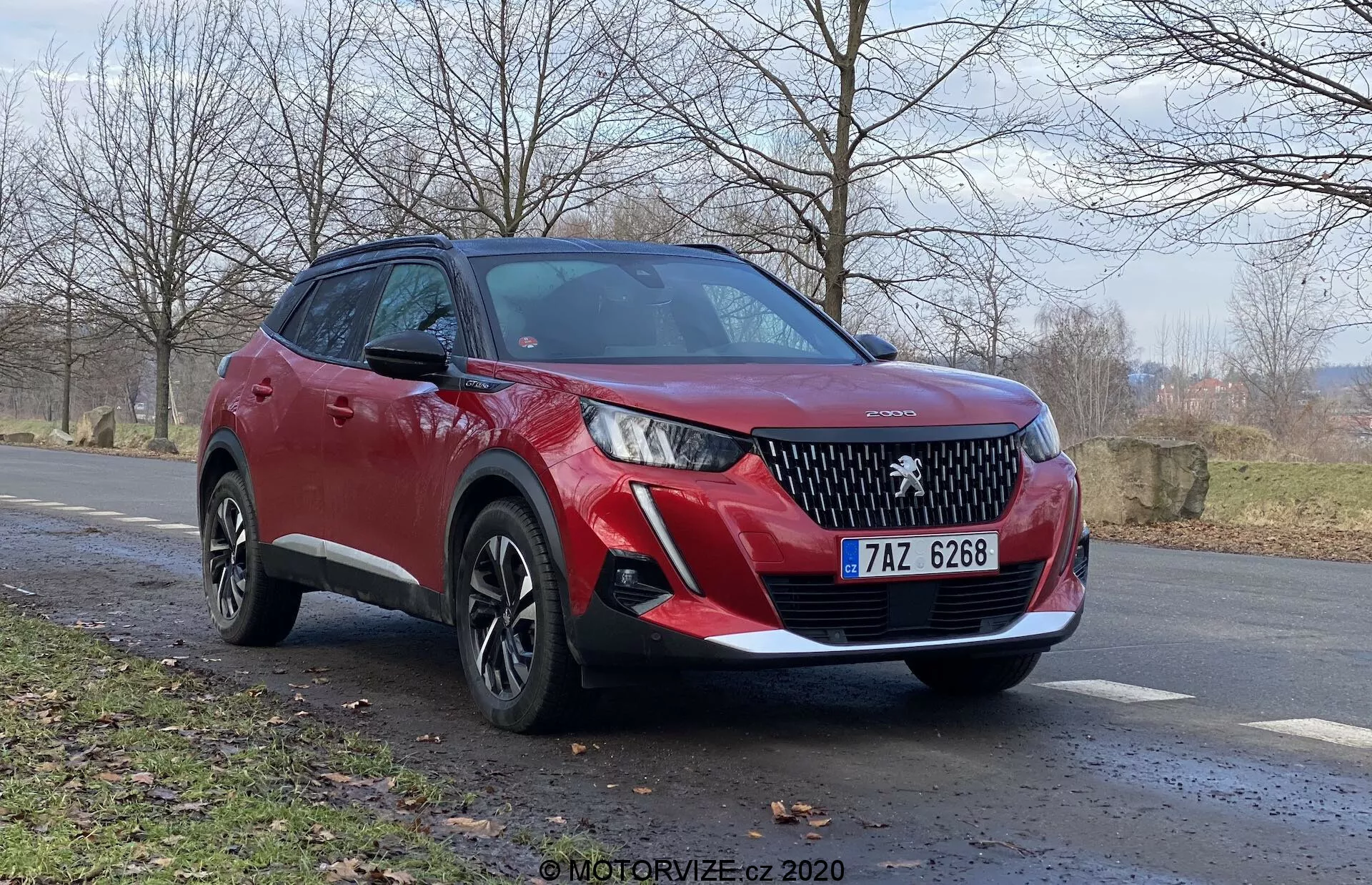 SUV rouge Peugeot 2008 2020 (Peugeot-2008-1-2-Puretech-100-Active) vu de trois quarts avant, mettant en avant la calandre avant avec un emblème de marque proéminent, des phares LED élégants, des capteurs de stationnement avant et des feux antibrouillard. La vue latérale révèle des roues en alliage à plusieurs rayons, une hauteur de conduite plus élevée, des lignes de carrosserie dynamiques, des clignotants intégrés dans les rétroviseurs latéraux et un revêtement en plastique noir. Positionné à un léger angle sur le côté de la route avec un paysage rural naturel à l'arrière-plan, indiquant la fin de l'automne ou le début du printemps. Angle de prise de vue bas pour une vue puissante et imposante.