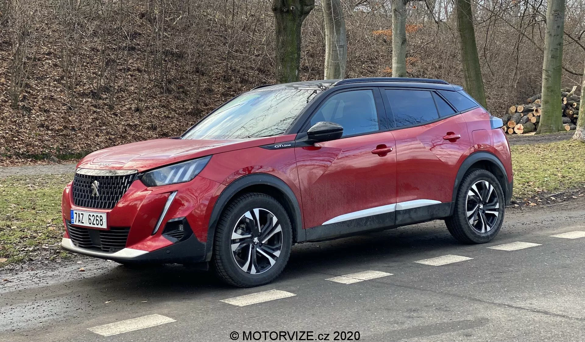 Perspective de côté et de face d'un crossover SUV moderne Peugeot 2008 1.2 PureTech 100 Active 2020, mettant en évidence sa calandre proéminente, ses phares élégants, ses roues en alliage et ses lignes dynamiques. Le véhicule, orné d'une finition métallique rouge avec vitres arrière teintées, est montré stationné sur une route latérale avec des rails de toit visibles, suggérant des capacités urbaines et légères tout-terrain. Image provenant de MOTORVIZE.cz 2020.