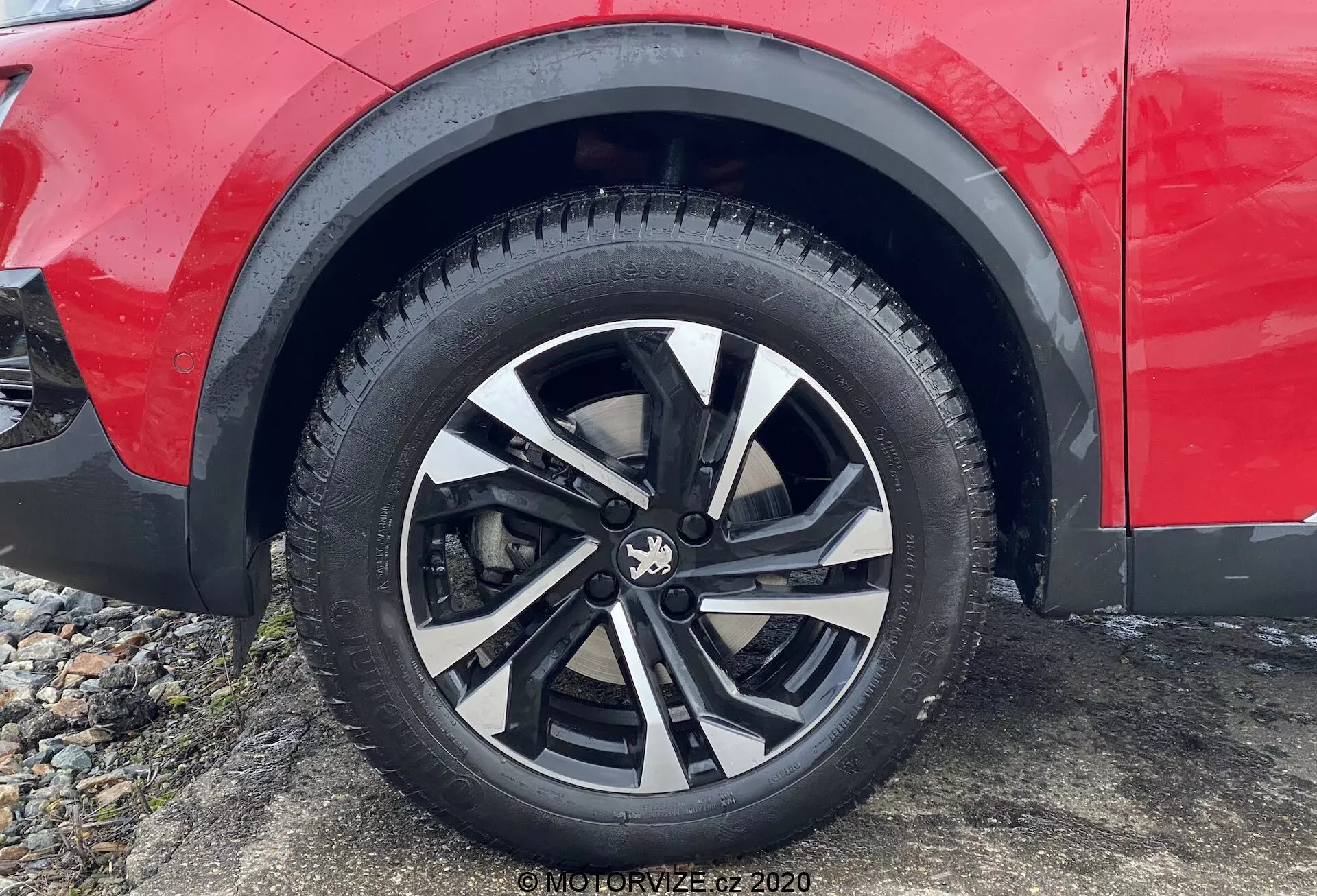 Gros plan d'une roue en alliage moderne au design multi-rayons avec des finitions noires et argentées, se concentrant sur la roue, le motif de la bande de roulement du pneu et une partie du panneau latéral de la voiture rouge près de l'aile de la roue d'un véhicule garé, suggérant un SUV ou un crossover adapté à la conduite sur route et hors route.