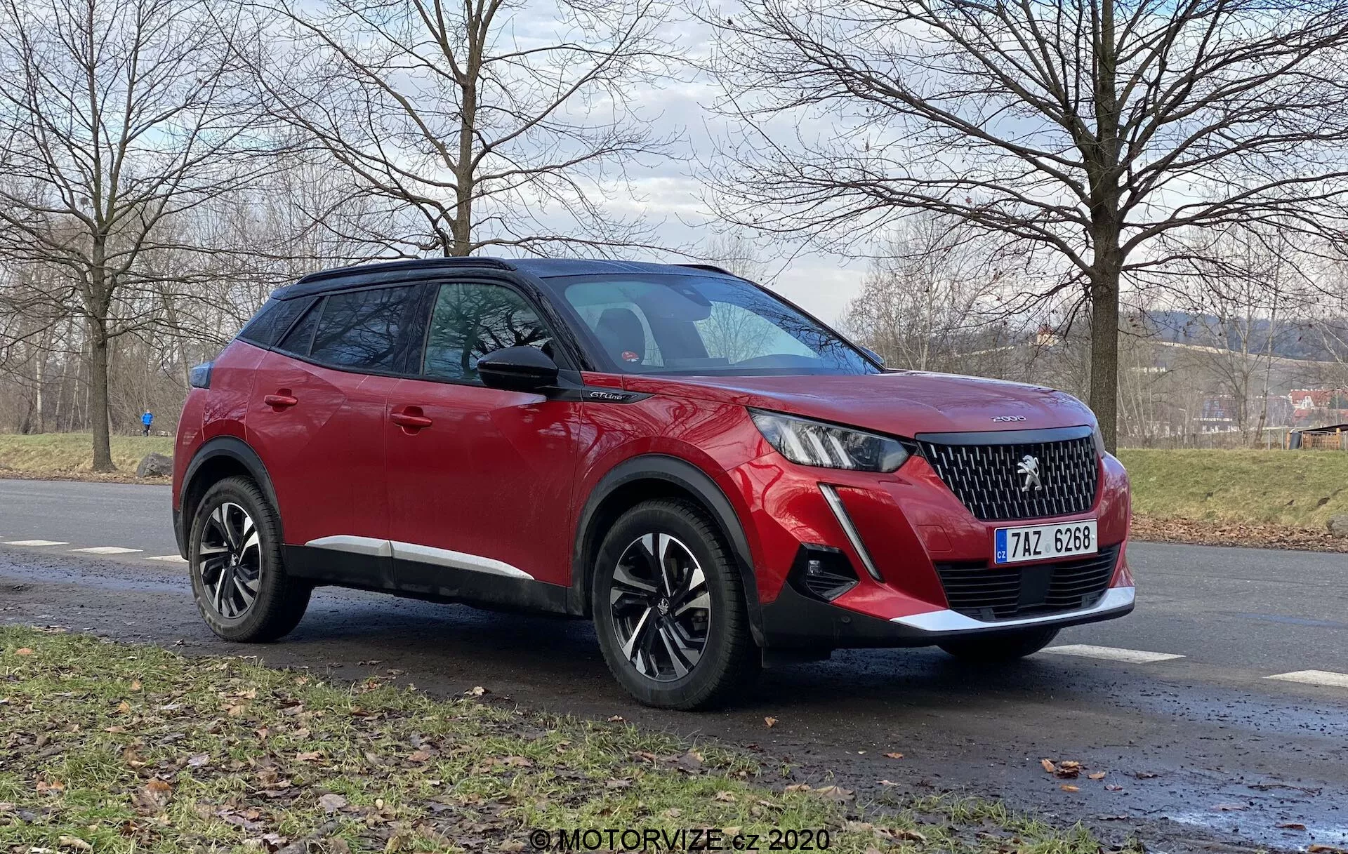 Vue de trois quarts avant d'un Peugeot 2008 2020 rouge (1.2 PureTech 100 Active) SUV crossover sur une route en asphalte. Les caractéristiques visibles incluent des feux de jour à LED, une calandre à motif diamant, un pare-chocs avant avec des feux antibrouillard et des capteurs de stationnement possibles, des roues en alliage à plusieurs branches et une caméra sous le rétroviseur latéral de la couleur de la carrosserie, le tout sous un ciel couvert. En arrière-plan, une zone herbeuse et des arbres sans feuilles, suggérant la fin de l'automne ou l'hiver.