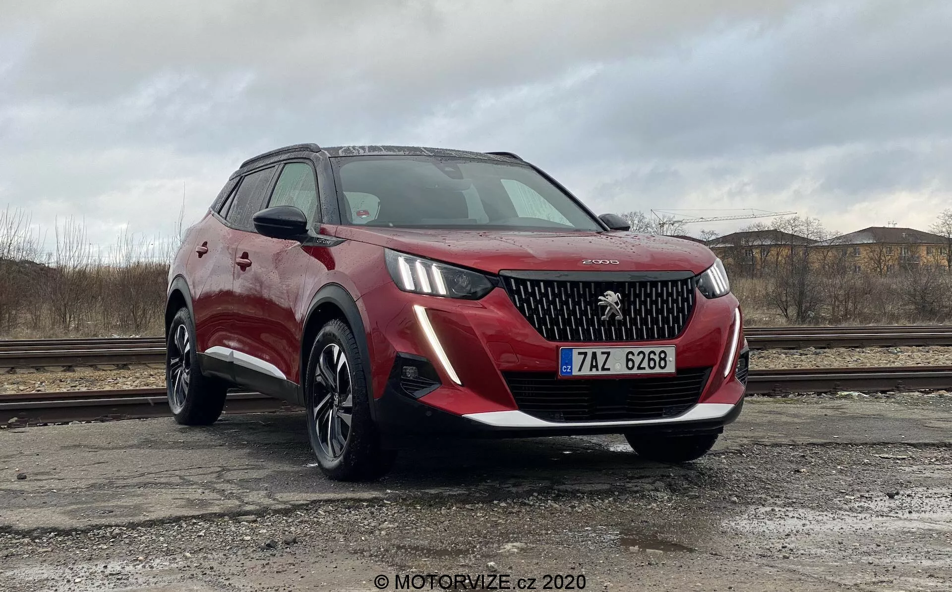 Vue de trois quarts avant d'une Peugeot 2008 1.2 PureTech 100 Active 2020 rouge, mettant en valeur sa grille détaillée, ses phares à LED, les lignes nettes du capot et les élégantes lumières diurnes. L'image capture le profil latéral côté conducteur, y compris le rétroviseur et la ligne de fenêtre, soulignant le design élégant et les caractéristiques technologiques avancées du véhicule. La voiture est probablement positionnée sur une surface en béton, avec des reflets et textures clairement visibles sur la carrosserie, offrant une perspective équilibrée adaptée aux objectifs marketing.