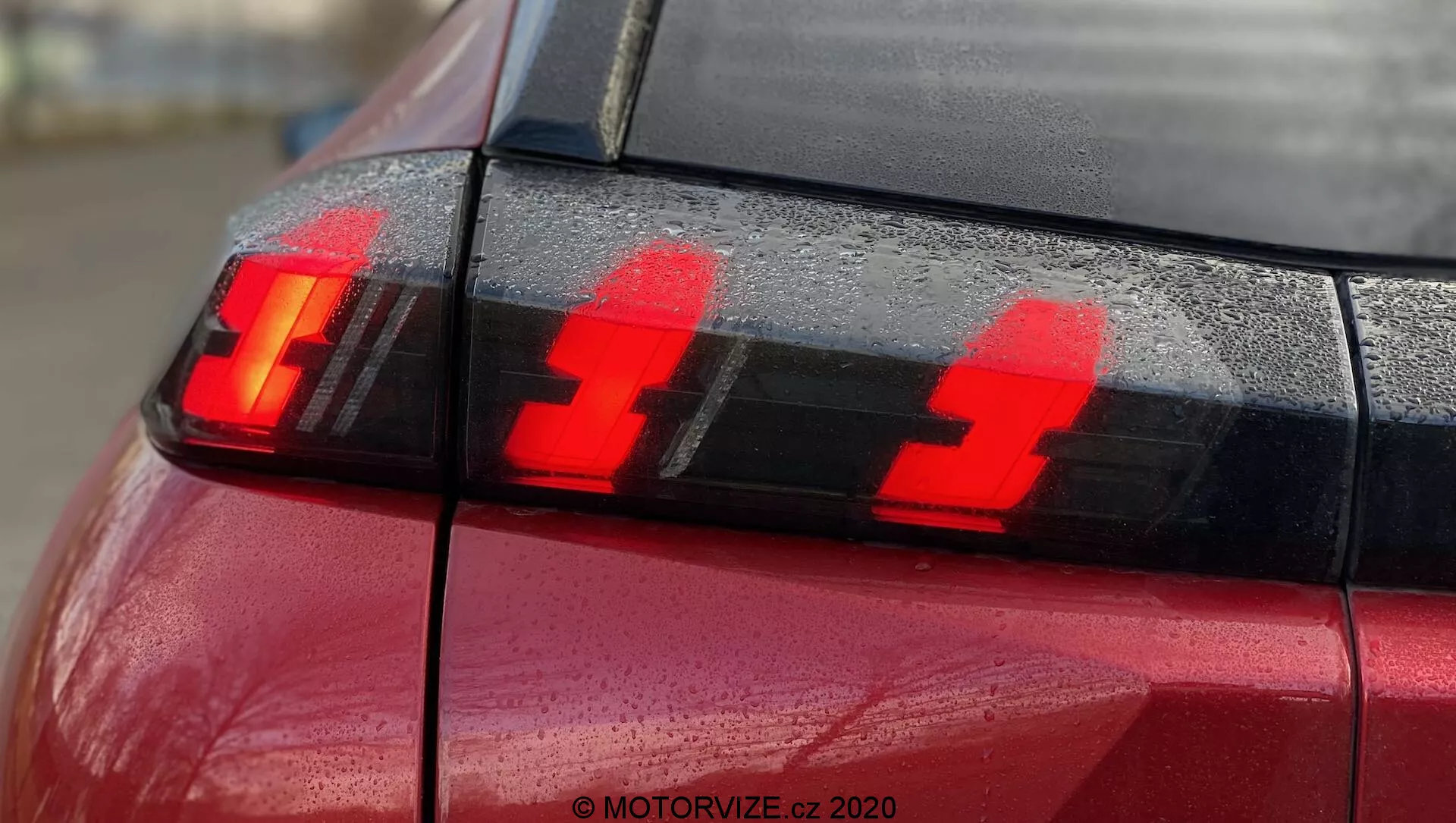 Gros plan de la section arrière d'une Peugeot 2008 2020 rouge (PureTech 100 Active) avec un focus sur l'ensemble des feux arrière montrant les feux de freinage allumés. La photographie, prise depuis l'angle arrière légèrement sur le côté, met en évidence trois éléments lumineux verticaux distincts à LED et des gouttelettes d'eau sur la surface, soulignant le design moderne de la technologie d'éclairage automobile.