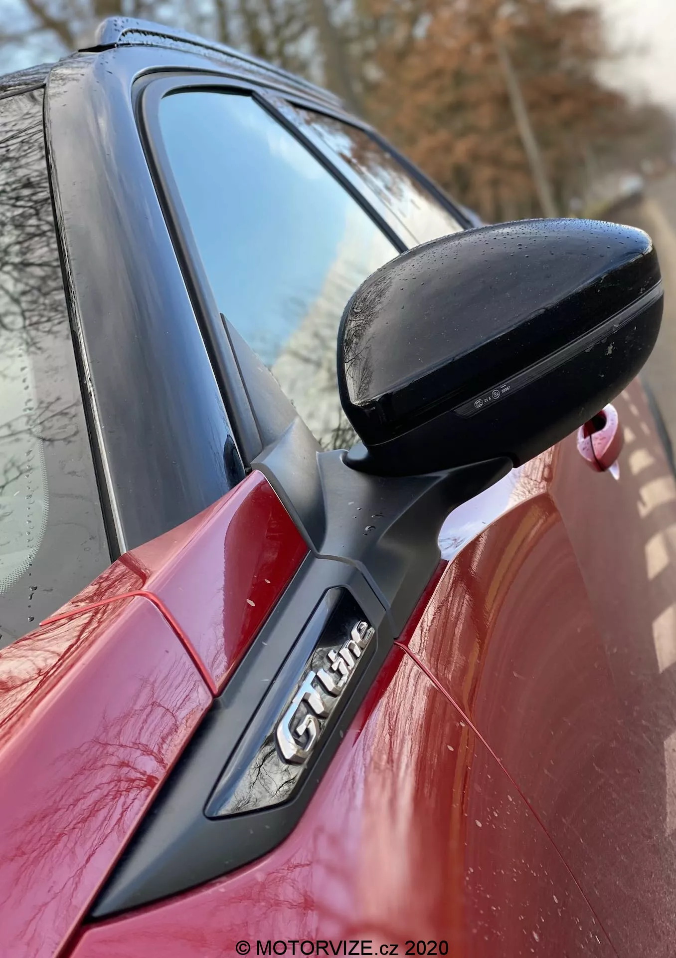 Gros plan sur le côté d'une Peugeot 2008 2020 rouge, mettant en avant le rétroviseur noir du côté conducteur, fixé à la porte avec un support noir. Le pilier A avec le logo 'GT Line' est partiellement visible, montrant les lignes de design aérodynamiques et les caractéristiques sportives de la voiture. Filigrane de MOTORVIZE.cz 2020.