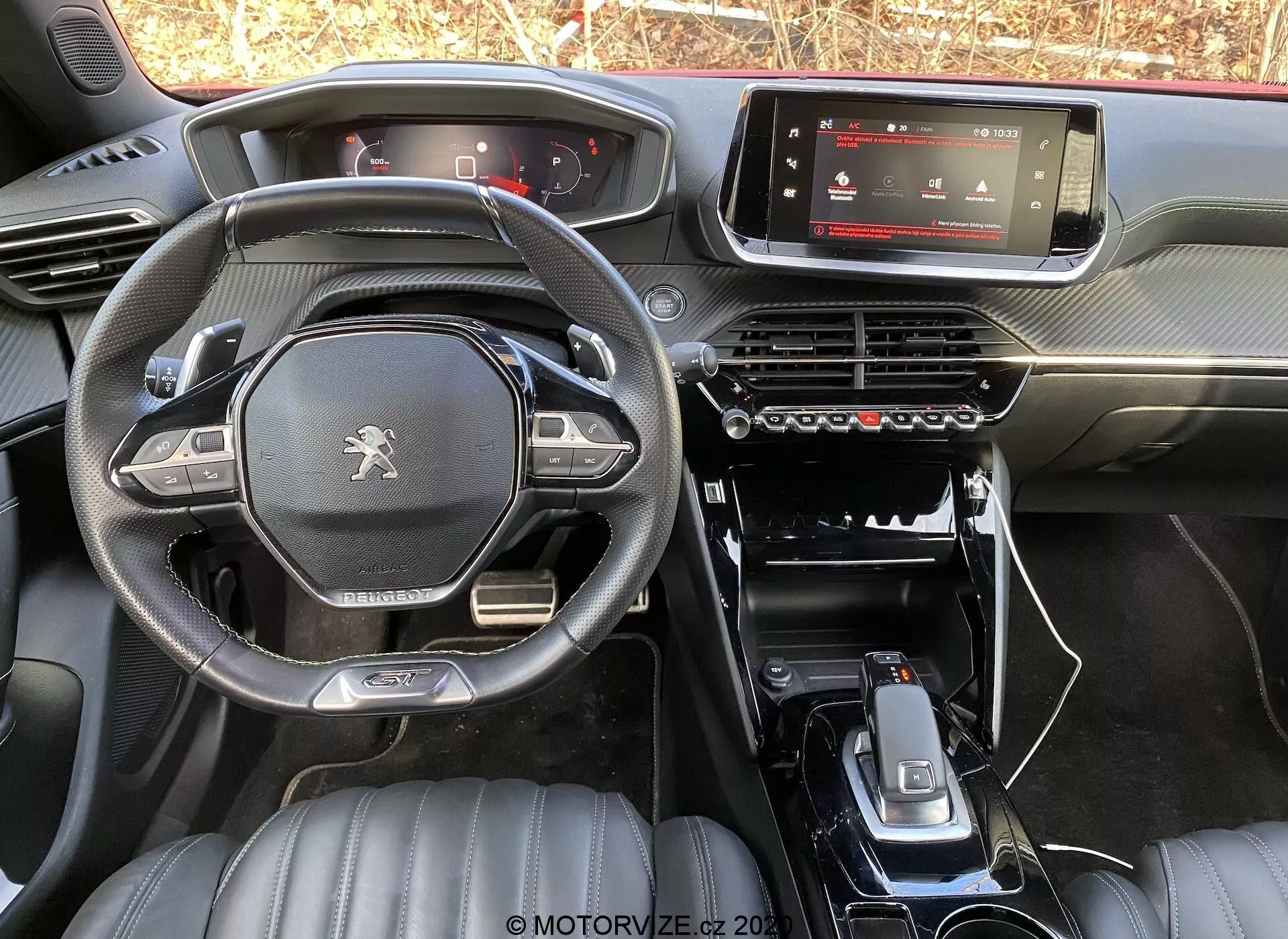 Vue intérieure de la Peugeot 2008 1.2 PureTech 100 Active 2020 depuis le siège arrière, montrant le tableau de bord, l'écran d'infodivertissement numérique, le panneau de contrôle de la climatisation, la console centrale minimaliste avec levier de vitesse, porte-gobelets et boutons de commande supplémentaires. Le volant à fond plat arbore le logo Peugeot et des commandes multifonctions. Les sièges avant en cuir avec motifs matelassés suggèrent un niveau de finition supérieur. L'éclairage naturel met en valeur le design et les matériaux du modèle d'usine.
