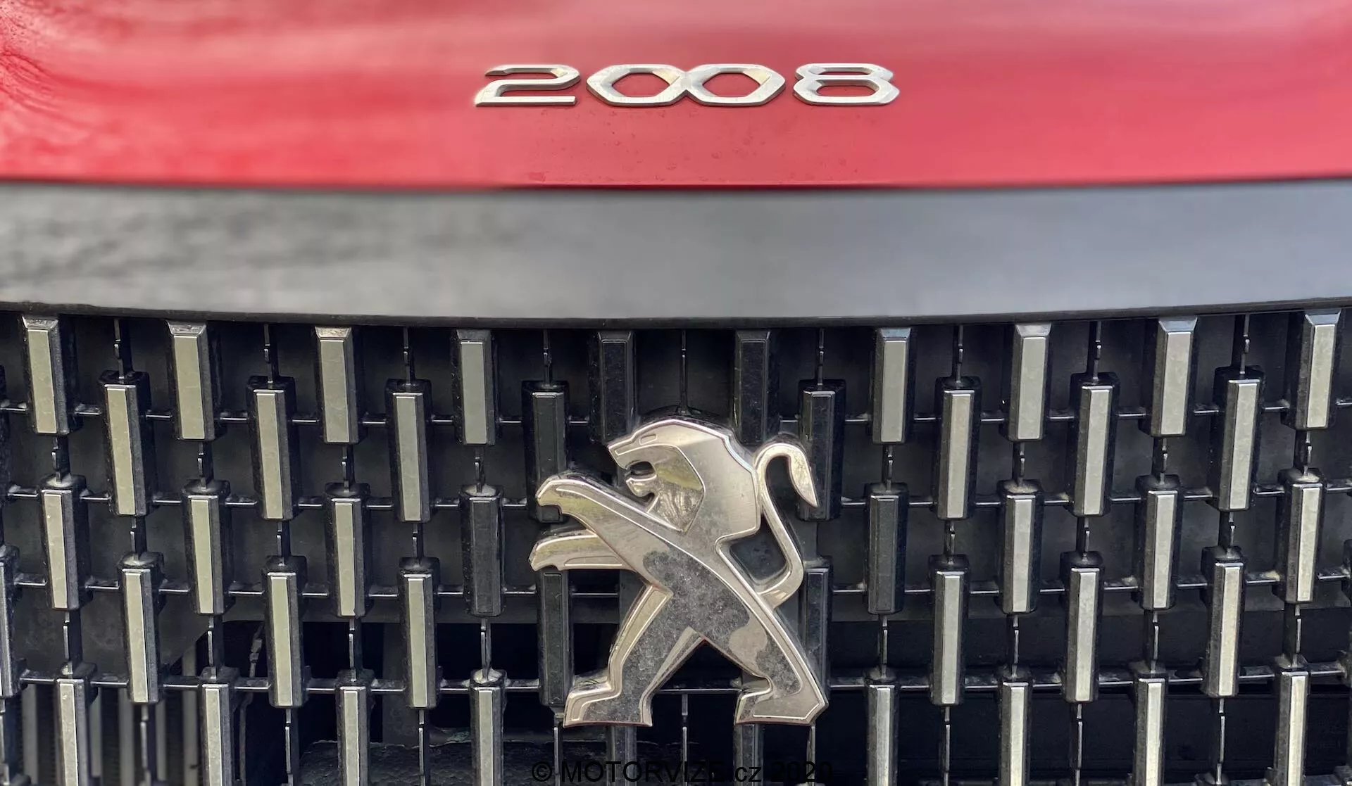 Vue en gros plan de la calandre avant d'une Peugeot 2008 1.2 PureTech 100 Active 2020, présentant le design distinctif avec un capot rouge vif, un ensemble complexe de lattes noires verticales et horizontales et un emblème de lion argenté au centre. La photo se concentre sur les éléments esthétiques et de marque, offrant une visualisation claire des détails de la calandre.