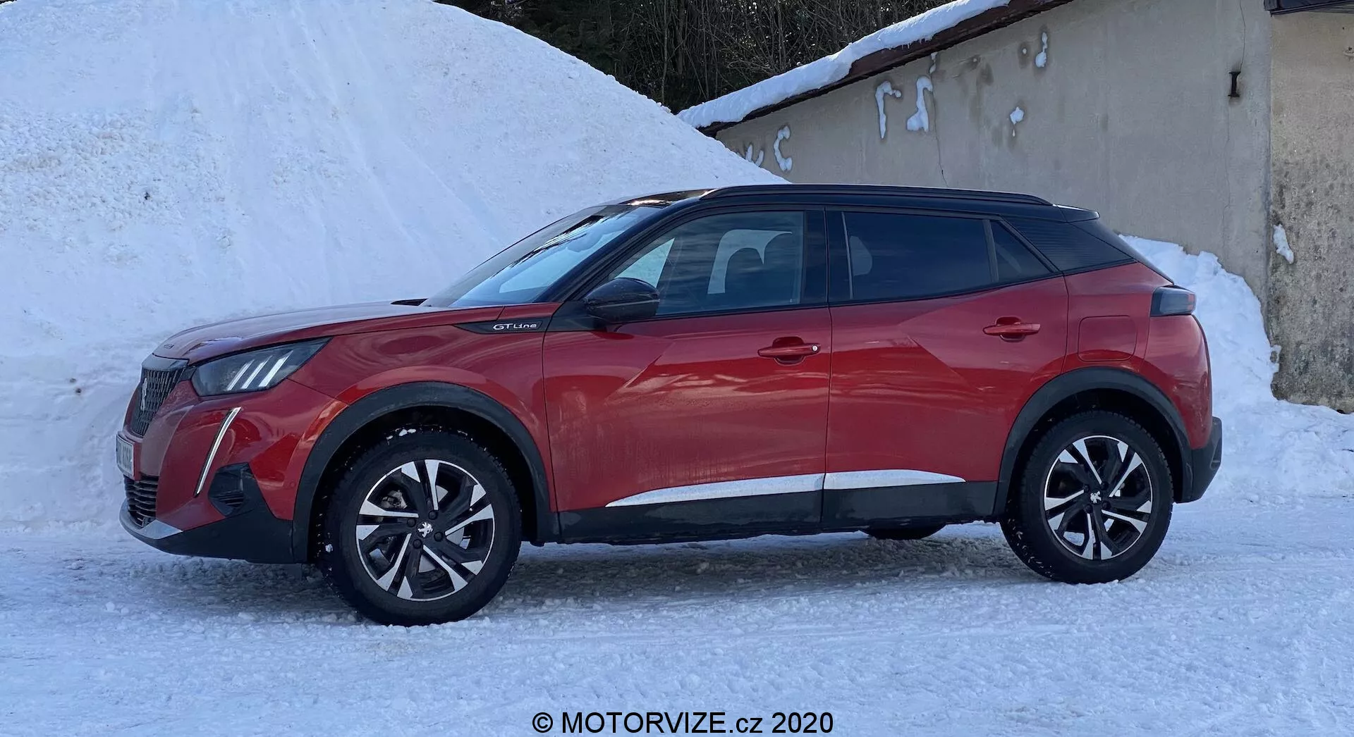 Vue de profil latéral d'un SUV crossover Peugeot 2008 2020 1.2 PureTech 100 Active à quatre portes rouge. Le véhicule est situé dans un environnement enneigé, soulignant ses capacités potentielles par temps froid. Il est équipé de roues en alliage à plusieurs rayons, de moulures en plastique noir le long du bas des portes et des arches de roue, et de fenêtres élégantes. Le design des phares et de la calandre se devine à l'avant, montrant un look moderne avec des phares LED probables. Les rétroviseurs latéraux comprennent des clignotants intégrés. Une ligne de toit en pente et un système de rails de toit suggèrent praticité et utilité pour un style de vie actif.