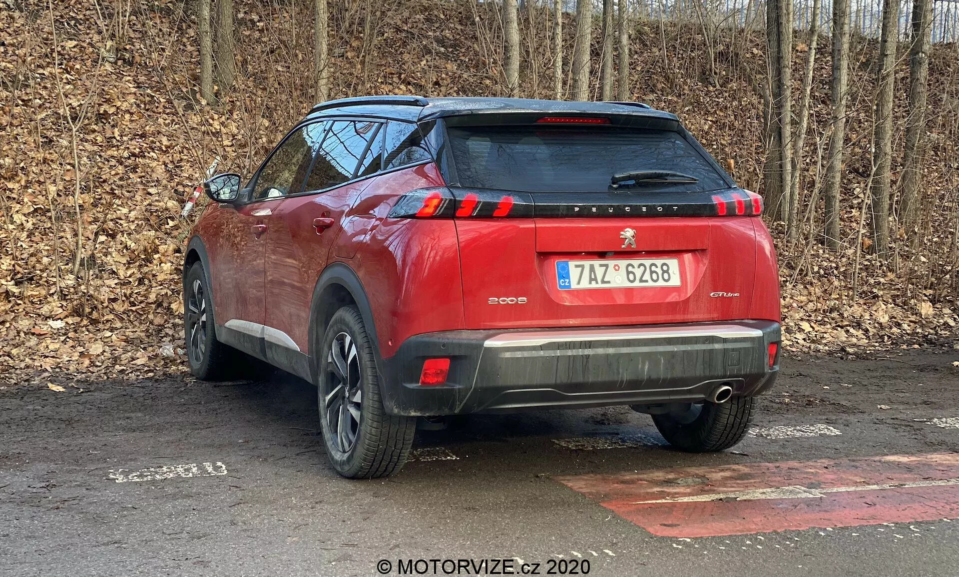 Vue arrière en angle d'un SUV Peugeot 2008 1.2 PureTech 100 Active rouge de 2020, garé sur une surface pavée, avec en toile de fond des arbres sans feuilles suggérant la fin de l'automne ou l'hiver. Capturée sous un angle arrière trois-quarts, l'image met en avant le côté droit du véhicule et montre des éléments de design tels que les feux arrière, la vitre arrière, le pare-chocs arrière bicolore avec des capteurs de stationnement, le becquet de toit, les bandes verticales des feux stop, les roues en alliage partiellement visibles et les barres de toit. L'échappement caché laisse présager un système de propulsion hybride ou électrique. Le véhicule occupe la majeure partie du cadre, indiquant la proximité du photographe.