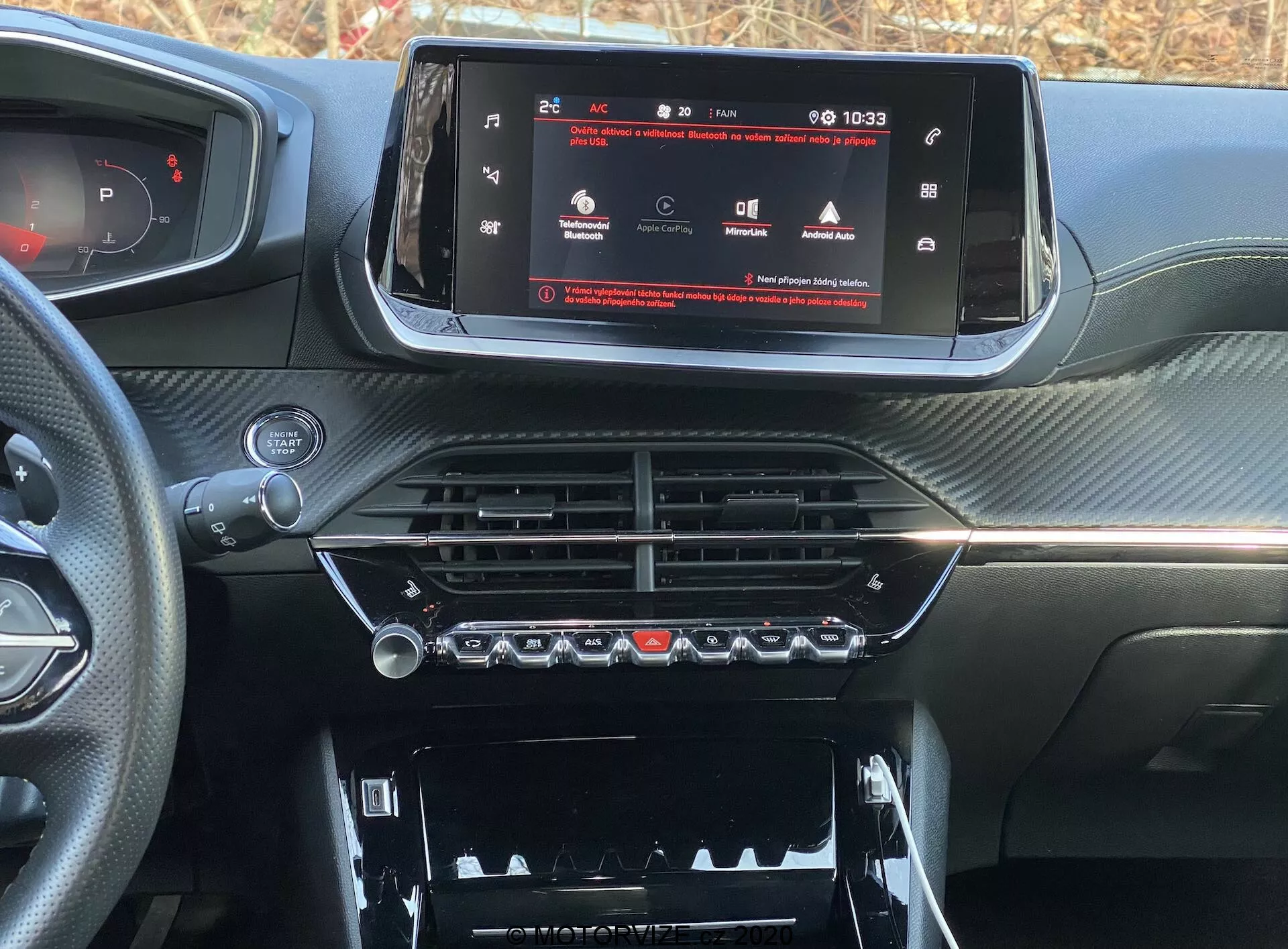 Vue rapprochée du tableau de bord intérieur d'une voiture moderne, spécifiquement la Peugeot 2008 2020 (peugeot-2008-1-2-puretech-100-active). L'image est prise du siège du conducteur ou du passager avant, se concentrant sur la console centrale et le système d'info-divertissement. Un écran tactile panoramique affiche un menu de réglages du véhicule, indiquant une technologie avancée. En dessous, des aérateurs chromés et des commandes climatiques avec des boutons physiques et des cadrans rotatifs. Plus bas, un compartiment avec un couvercle est visible. Le tableau de bord a une finition texturée semblable à la fibre de carbone, donnant un aspect sportif et haut de gamme.