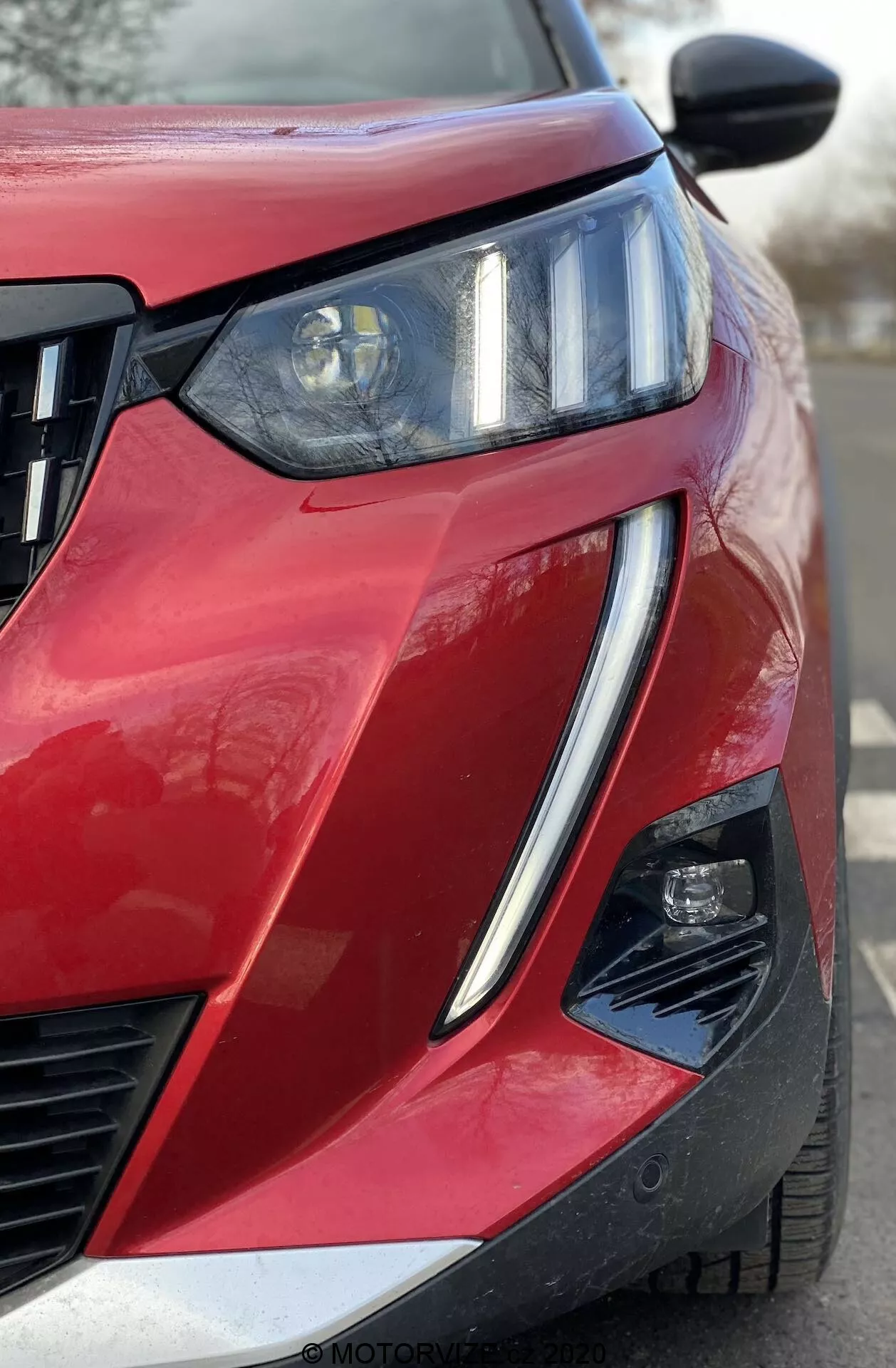 Vue frontale rapprochée et angulaire d'une Peugeot 2008 1.2 PureTech 100 Active rouge de 2020, mettant en avant le quart avant côté conducteur. Phare proéminent avec faisceau projecteur et DRL à LED intégrés, grille à lattes horizontales et assemblage secondaire de lumière partiellement visible dans une découpe angulaire du pare-chocs. Peinture brillante avec finition métallique, ligne de carrosserie nette, contour surélevé du capot, rétroviseur latéral noir avec potentiellement des clignotants intégrés ou une caméra d'angle mort, et découpe dans le coin inférieur du pare-chocs suggérant des systèmes avancés d'assistance à la conduite.