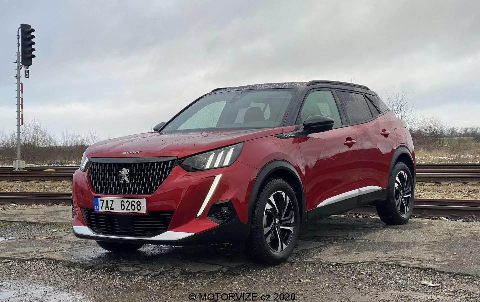 Vue de trois quarts avant d'un SUV crossover Peugeot 2008 1.2 PureTech 100 Active rouge moderne. La prise de vue en angle bas met en valeur la hauteur et la posture du véhicule. L'avant présente une calandre proéminente avec un motif en losange, un emblème représentant probablement la marque du constructeur et des phares distinctifs intégrant des feux diurnes à LED. Le pare-chocs avant contient des antibrouillards ou des entrées d'air. Le profil latéral révèle des lignes aérodynamiques, de grandes roues en alliage à plusieurs branches, des barres de toit et des rétroviseurs avec clignotants intégrés. L'image capture le design et les caractéristiques technologiques du SUV par un jour nuageux, garé près de voies ferrées.