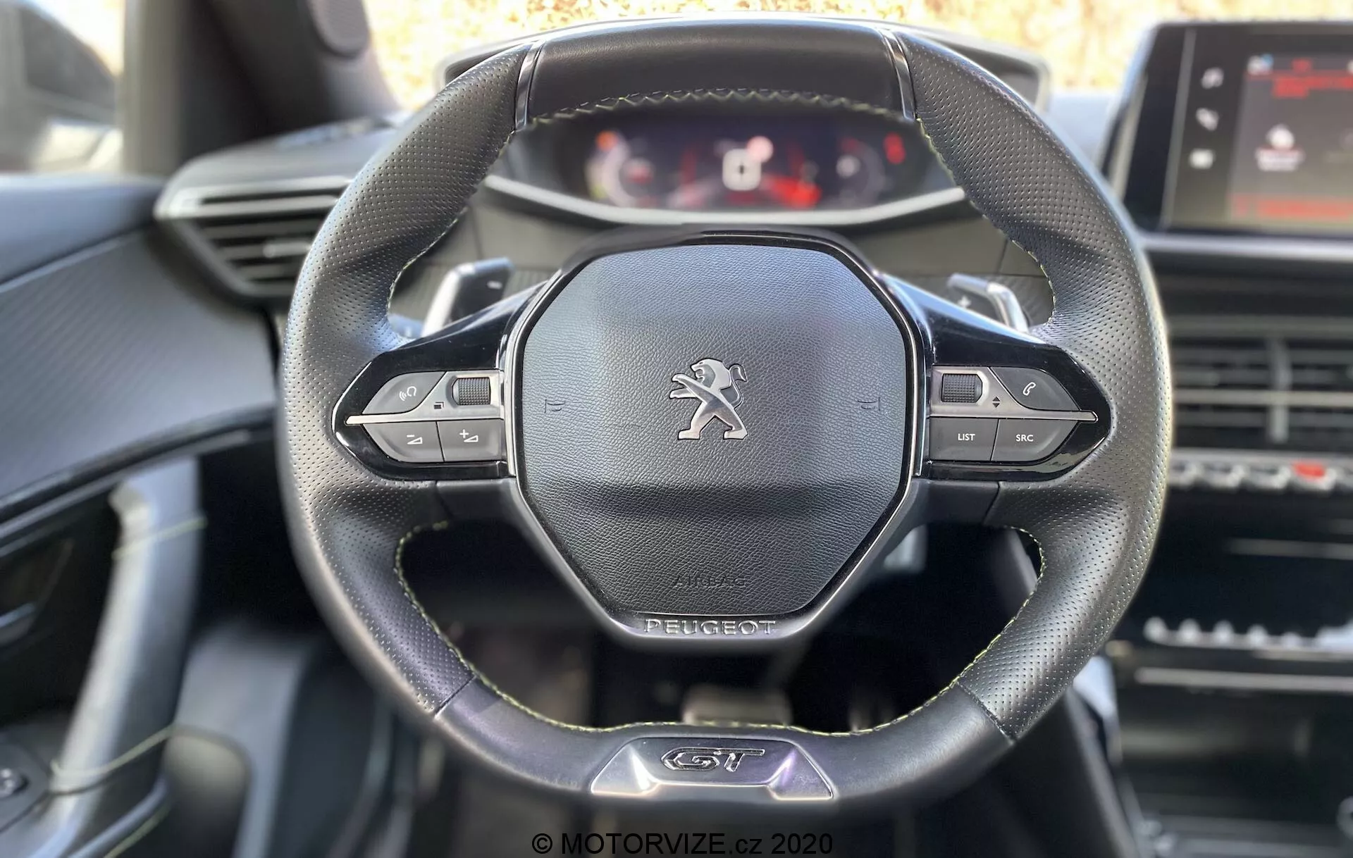 Vue intérieure de la Peugeot 2008 2020 centrée sur le volant et le tableau de bord depuis la perspective du conducteur. Comprend un volant en cuir à trois branches avec boutons multifonctions, l'emblème du lion Peugeot et un tableau de bord numérique personnalisable. Eléments visibles incluent écran tactile d'infodivertissement, aérateurs et contrôles de la console centrale avec un éclairage naturel adoucissant les détails du volant.