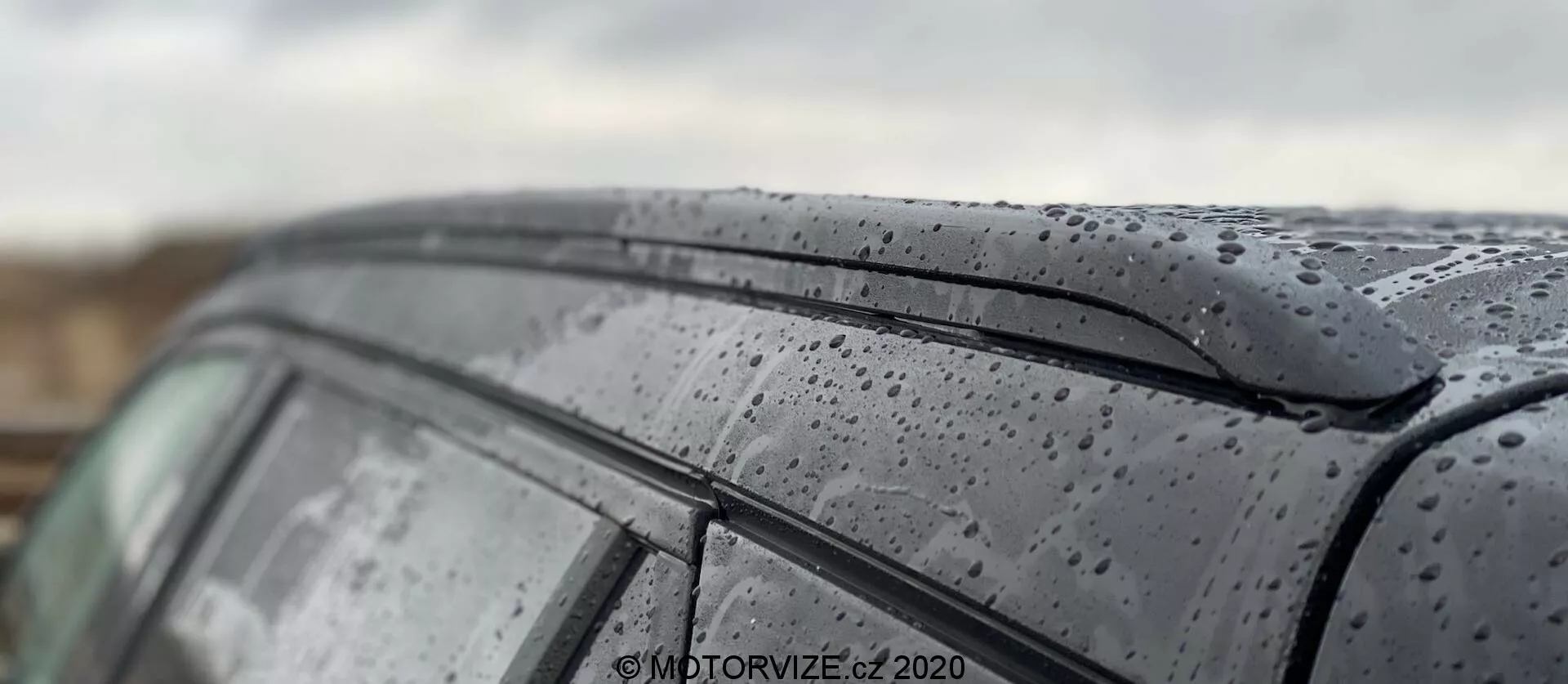 Gros plan sur le toit et la vitre latérale du modèle Peugeot 2008 1.2 PureTech 100 Active 2020, mettant en évidence les gouttes de pluie sur la surface élégante. L’image capture la peinture foncée et brillante avec les gouttes de pluie et le design minimaliste du bord supérieur de la vitre latérale. La faible profondeur de champ accentue la texture extérieure de la voiture tout en floutant l’arrière-plan et certaines parties du toit, montrant une représentation artistique de la voiture en conditions humides.