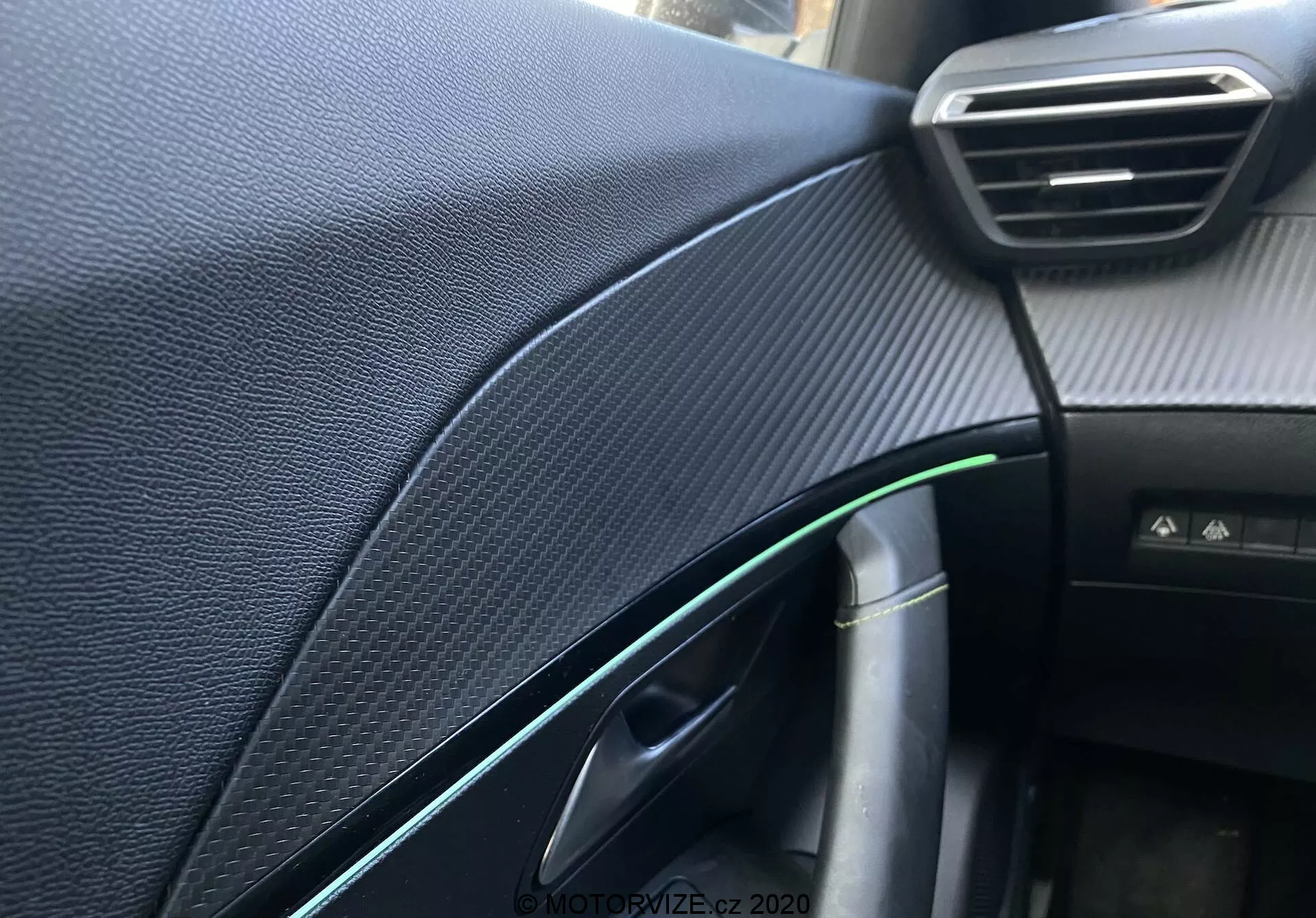 Vue rapprochée de l'intérieur du tableau de bord et des détails du côté passager de la Peugeot 2008 2020, se concentrant sur le tableau de bord synthétique texturé noir ressemblant à la fibre de carbone, la ventilation réglable visible, la porte partiellement ouverte avec un reflet vert et un accent chromé, la poignée de porte, un câble avec connecteur de charge, la lumière de cabine et un bouton non identifié, capturés avec une faible profondeur de champ et un éclairage naturel.