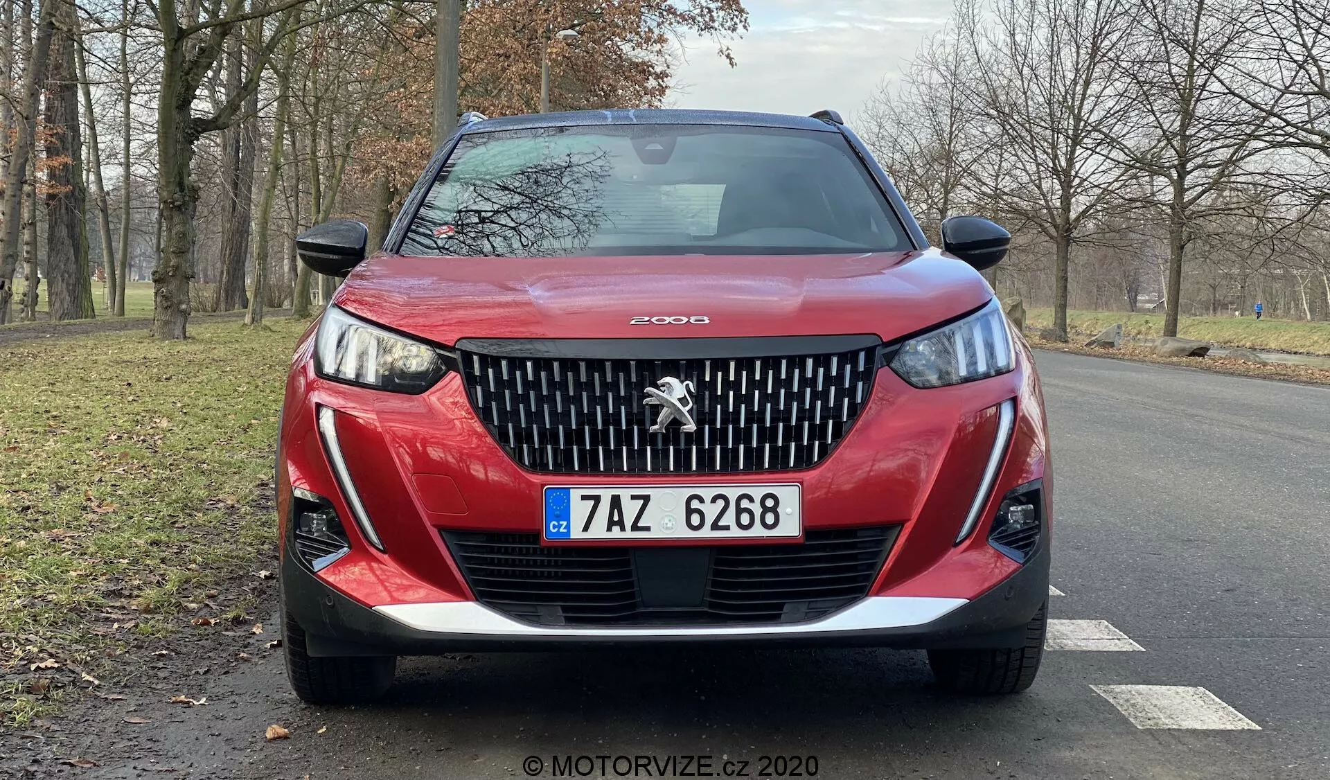 Vue frontale rapprochée d'une Peugeot 2008 2020 rouge (Peugeot-2008-1-2-PureTech-100-Active) mettant en valeur sa calandre détaillée avec l'éminent emblème du lion Peugeot, le design agressif des phares avec des feux de jour à LED en forme de 'crocs de sabre', un pare-chocs inférieur sculpté et une plaque d'immatriculation tchèque. Le contour du capot ajoute au dynamisme et l'image capture une vue partielle des jantes en alliage et des rétroviseurs sur une route asphaltée sèche avec des arbres sans feuilles et une zone herbeuse en arrière-plan, suggérant un environnement naturel et serein, possiblement en automne ou en hiver.