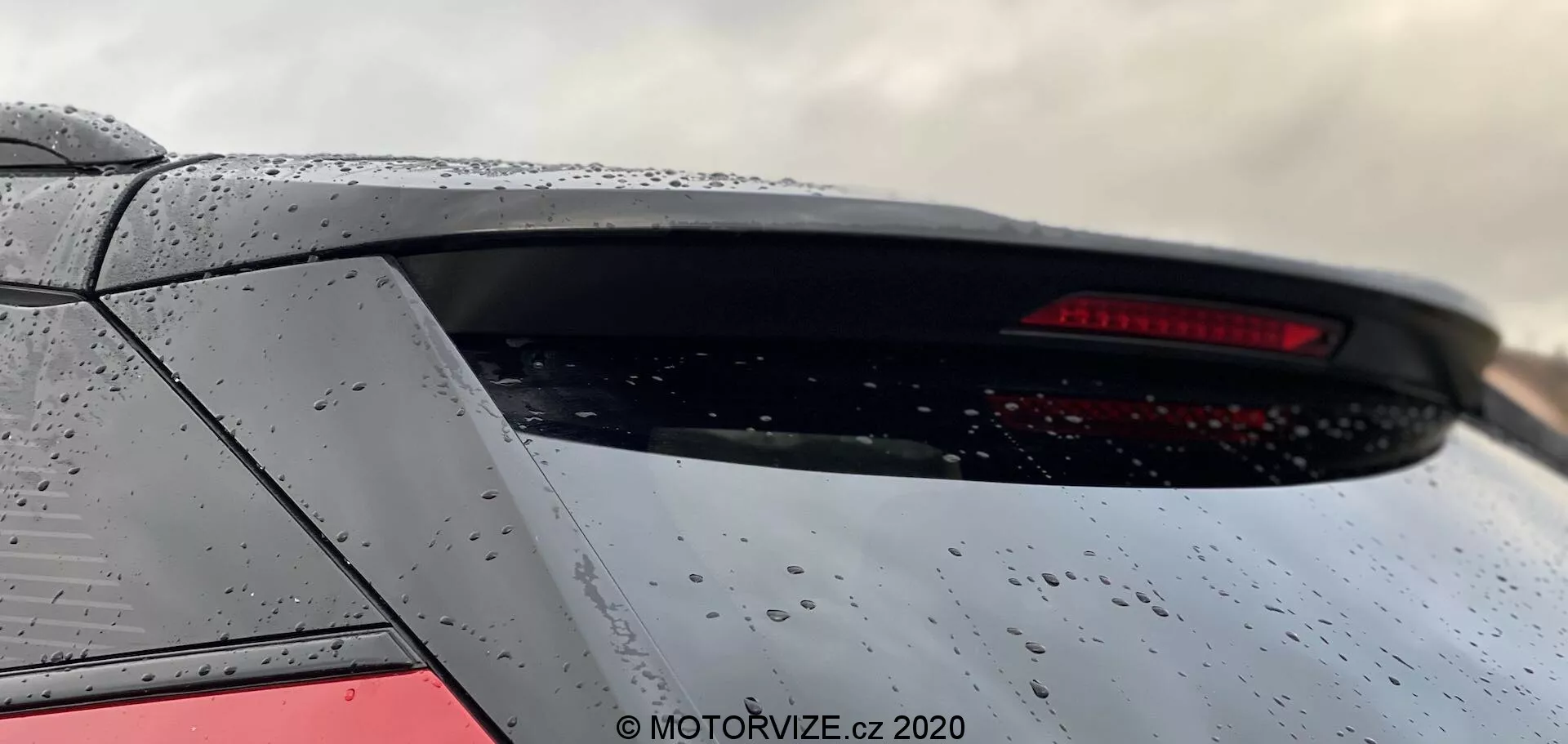 Vue rapprochée du segment arrière d'un modèle Peugeot 2008 2020 mettant en évidence la partie inférieure de la lunette arrière et la partie supérieure du coffre. L'angle ascendant depuis le niveau du sol met en avant la lunette arrière de la voiture, le spoiler intégré et le feu arrière LED. La voiture gris métallisé affiche des gouttes d'eau suggérant une pluie récente. L'image recadrée ne montre pas de logos identifiables.