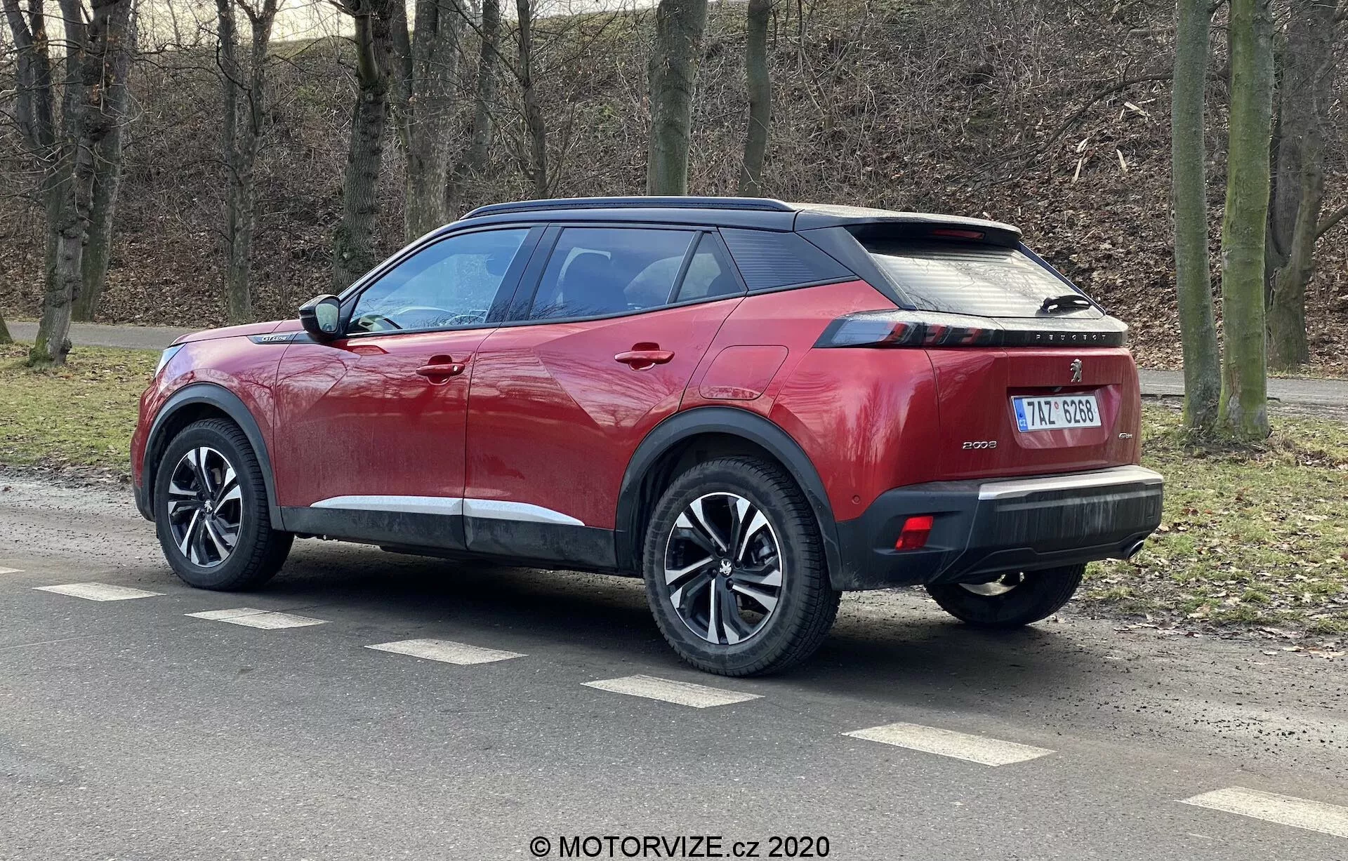 L'image montre un SUV Peugeot 2008 2020 rouge métallisé brillant avec un toit noir contrastant, des roues à plusieurs branches et des pneus à profil bas. Photographie prise d'une vue arrière trois quarts, le SUV affiche ses lignes latérales nettes, son spoiler intégré, une antenne en forme de nageoire de requin, des feux arrière LED horizontaux étroits et un pare-chocs arrière élégant avec des capteurs de stationnement. Le véhicule possède des poignées de porte et des rétroviseurs latéraux couleur carrosserie avec des clignotants intégrés, des vitres arrière teintées et une plaque d'immatriculation centrale sous un emplacement potentiel pour une caméra arrière.