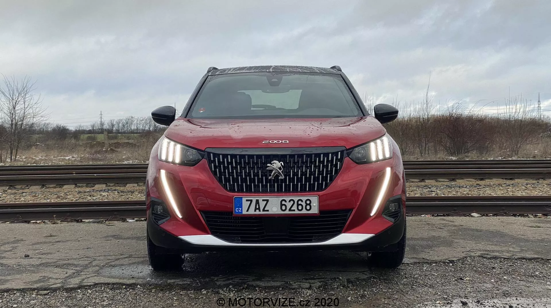 Vue frontale d'une Peugeot 2008 2020 PureTech 100 Active rouge, photographiée sous un angle bas mettant en valeur la calandre avant, le pare-chocs, les phares et l'emblème Peugeot. La voiture est propre avec des gouttes d'eau sur le capot, pare-brise clair reflétant un ciel couvert. Les feux de stationnement sont allumés, la plaque d'immatriculation européenne est visible. Le décor comprend un sol mouillé, des voies ferrées, des arbres dénudés et un ciel gris, créant une atmosphère industrielle.