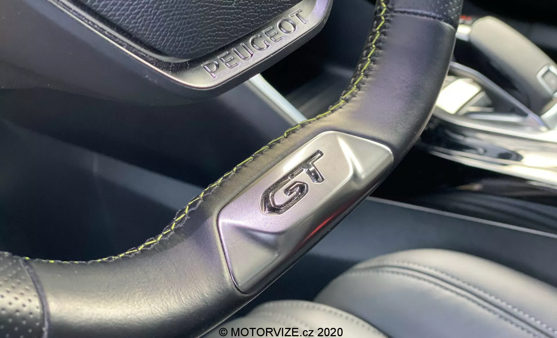 Gros plan de l'intérieur du Peugeot 2008 2020, mettant en avant le badge 'GT' sur le volant en cuir perforé avec des coutures vertes, soulignant sportivité et luxe. L'image se concentre sur les détails de l'artisanat et l'arrière-plan flou révèle des matériaux intérieurs premium et un design moderne.