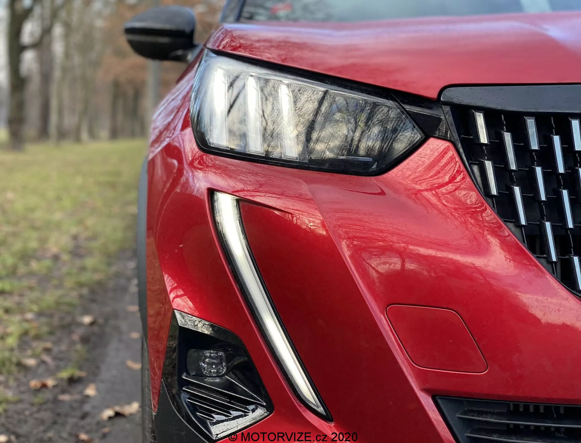 Vue rapprochée en contre-plongée de la section avant-gauche de la Peugeot 2008 2020 avec une finition rouge brillante. L'image met en évidence le phare LED angulaire et élégant, le design aiguisé de la prise d'air et les lignes sculptées, reflétant une technologie d'éclairage avancée et une esthétique aérodynamique.
