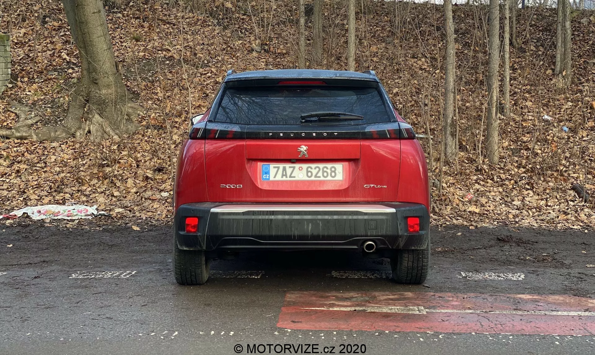 Vue arrière d'une voiture rouge garée à l'extérieur avec en arrière-plan des buissons ou des arbres sans feuilles, suggérant l'automne ou l'hiver. Les caractéristiques de la voiture incluent un logo central et une plaque d'identification sur le couvercle du coffre, une plaque d'immatriculation, un essuie-glace arrière, un spoiler arrière, des feux arrière modernes, des réflecteurs et des capteurs de stationnement arrière sur le pare-chocs, un pare-chocs arrière proéminent avec un tuyau d'échappement visible, une plaque de protection arrière, une antenne en forme de nageoire de requin sur le toit et un autocollant pelé sur le bas-gauche du couvercle du coffre.