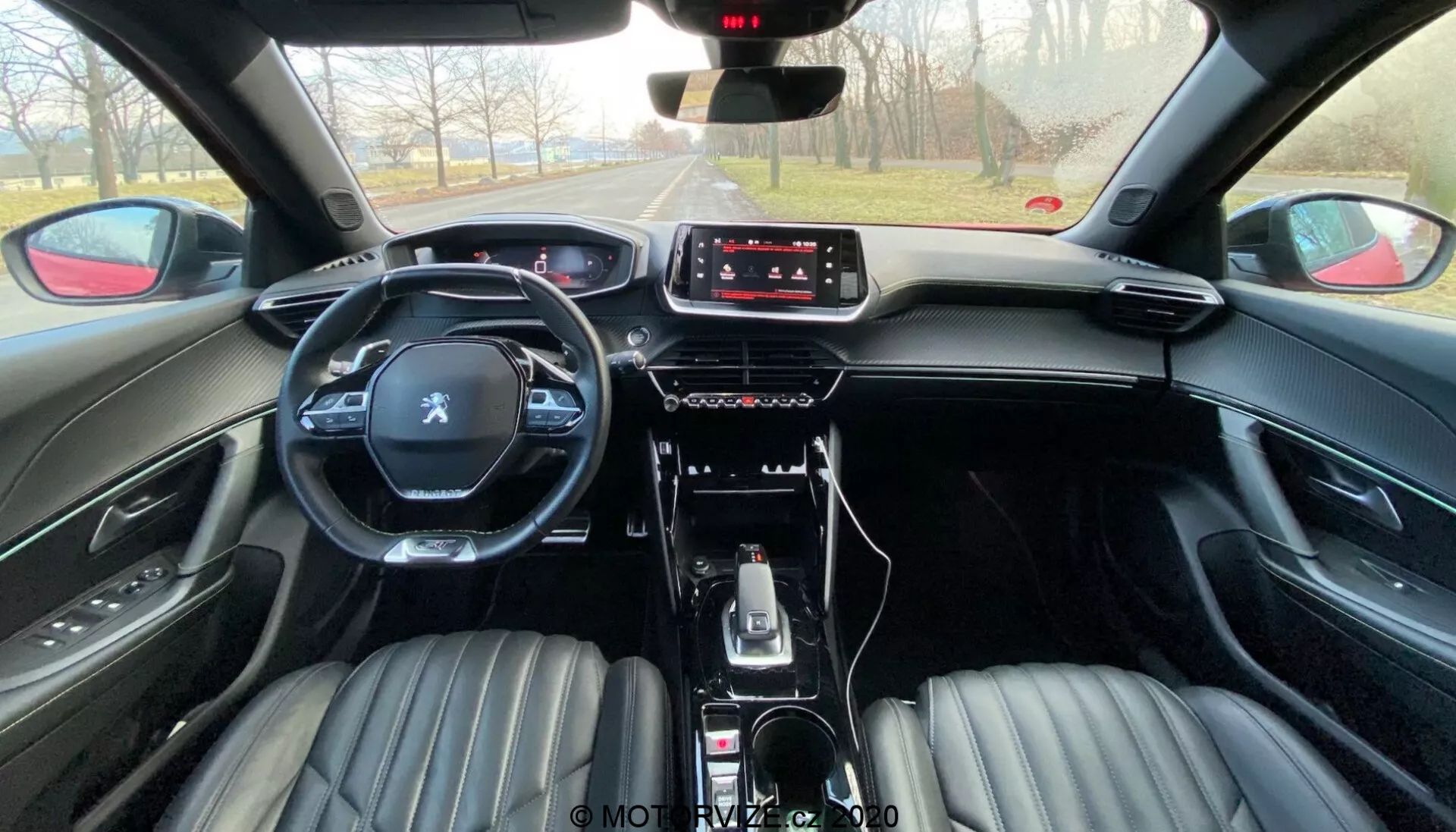 Vue intérieure du Peugeot 2008 2020 (peugeot-2008-1-2-puretech-100-active) depuis la perspective de la banquette arrière. L'image montre des sièges avant revêtus de cuir noir avec un design côtelé, des appuie-têtes intégrés, un volant multifonction avec le logo Peugeot et une console centrale moderne. Le tableau de bord est équipé d'un écran tactile d'infodivertissement avec des commandes physiques minimales, indiquant une interaction axée sur le tactile. La voiture comprend un levier de vitesses automatique, des sélecteurs de modes de conduite, un frein de parking électronique et des compartiments de rangement. L'atmosphère suggère une journée ensoleillée avec une vue dégagée sur la route à venir.