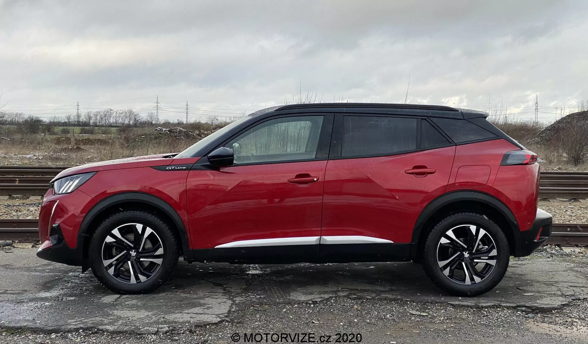 Profil latéral d'un Peugeot 2008 2020 SUV rouge vif, modèle 1.2 PureTech 100 Active, stationné sur une surface de gravier avec une végétation clairsemée et un ciel nuageux en arrière-plan. L'image montre le véhicule avec de grandes roues en alliage noir à plusieurs branches, un design contemporain et élégant avec une haute ceinture de caisse, quatre poignées de porte, des vitres teintées, des rétroviseurs latéraux saillants avec clignotants intégrés et un design de toit flottant avec des montants arrière noirs. Une ligne de garniture court le long de la partie inférieure de la voiture.