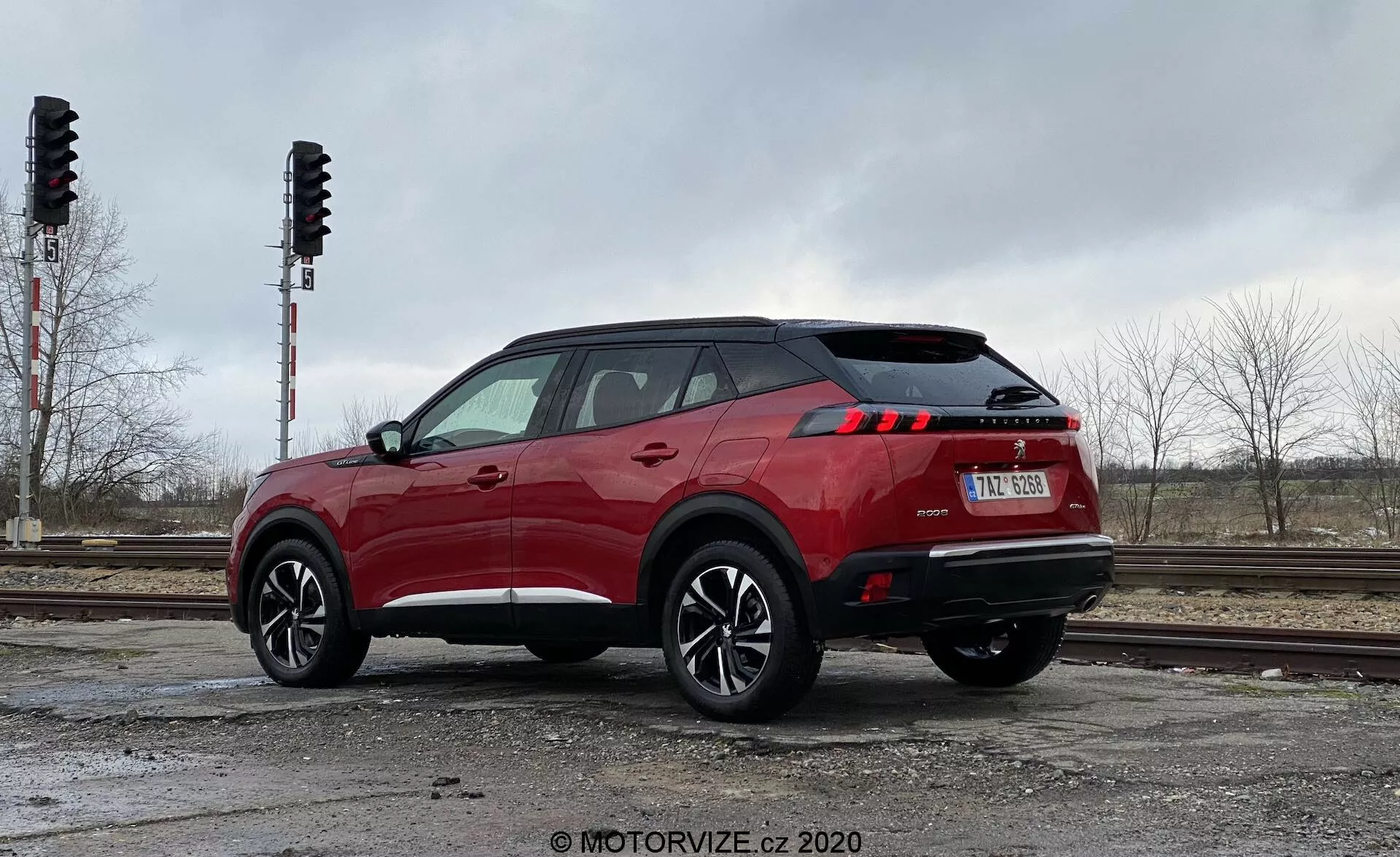 L'image présente un SUV moderne, spécifiquement le Peugeot 2008 2020 (1.2 PureTech 100 Active), capturé d'une vue arrière trois quarts. L'angle offre une perspective claire du profil latéral et arrière du véhicule, mettant en valeur son design et ses dimensions. Positionné à gauche avec amplement d'espace à droite, la voiture semble prête à bouger. La prise de vue est réalisée à partir d'un angle bas, près du sol, légèrement vers le haut, accentuant la hauteur et la posture du véhicule. La roue arrière et l'aile sont proéminentes, montrant le design de la roue et la hauteur de la suspension. La couleur rouge vif de la carrosserie contraste avec l'arrière-plan terne. La lumière diffuse d'une journée nuageuse offre une visibilité nette des détails, y compris les feux arrière à LED. L'environnement comprend des voies ferrées et des arbres, indiquant un cadre urbain ou industriel.