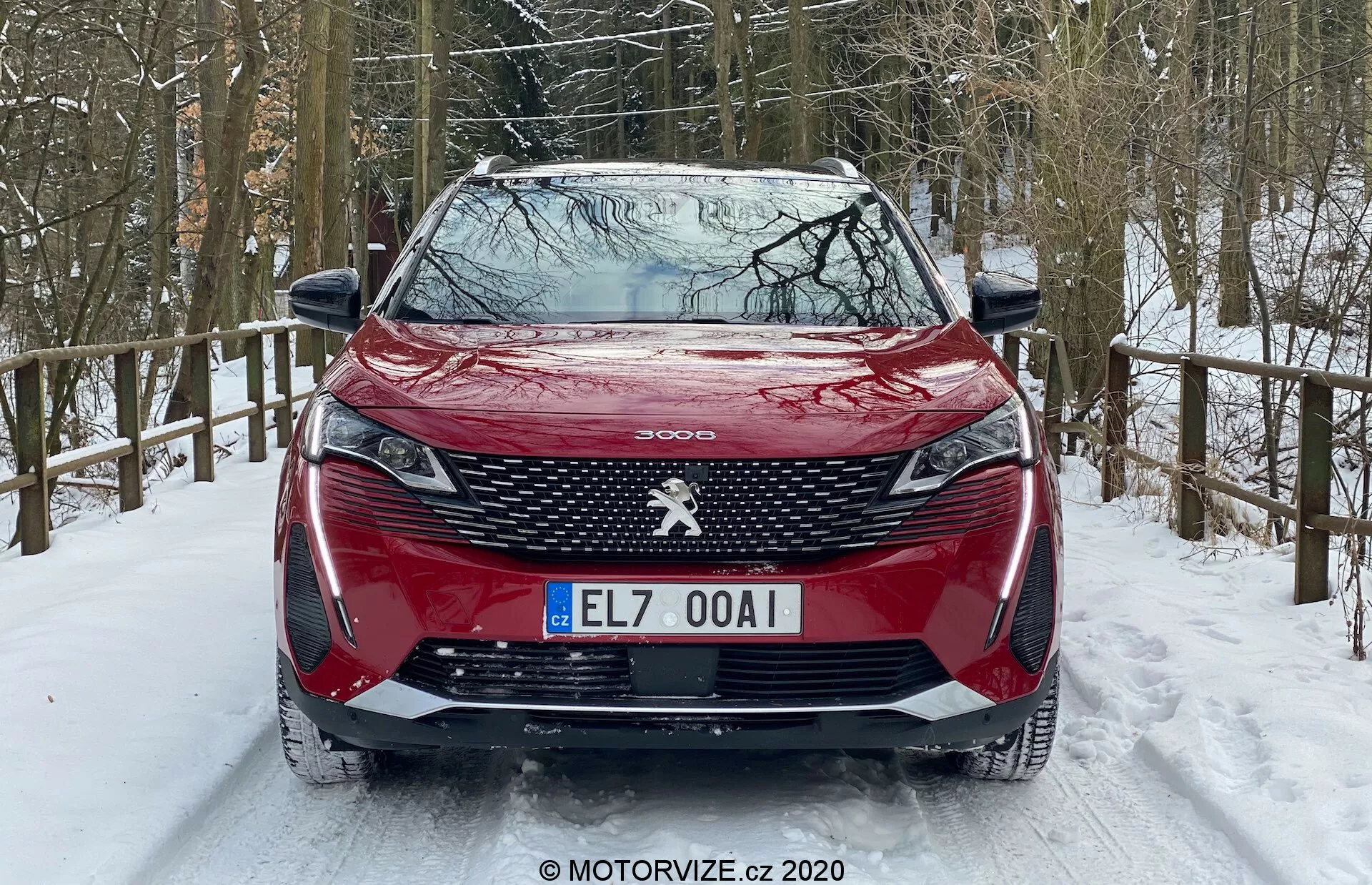 Vue avant d'une Peugeot 3008 rouge 2021 stationnée sur une route enneigée, avec motif de calandre distinctif, emblème Peugeot central, phares avant acérés avec feux diurnes en forme de crocs, capot profilé, pare-chocs avant avec capteurs de stationnement intégrés et environnement forestier enneigé.